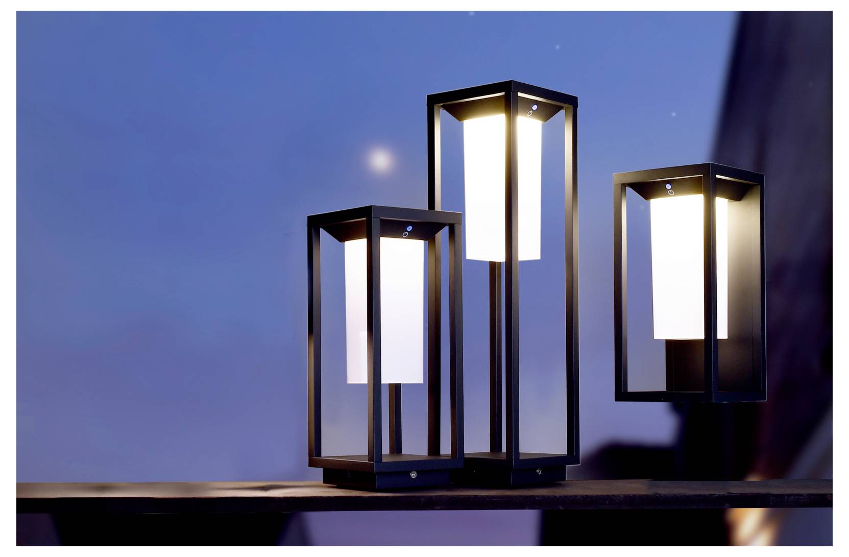 Tre lampade da tavolo a forma di lanterna luminose su una superficie scura, su uno sfondo di sera blu.