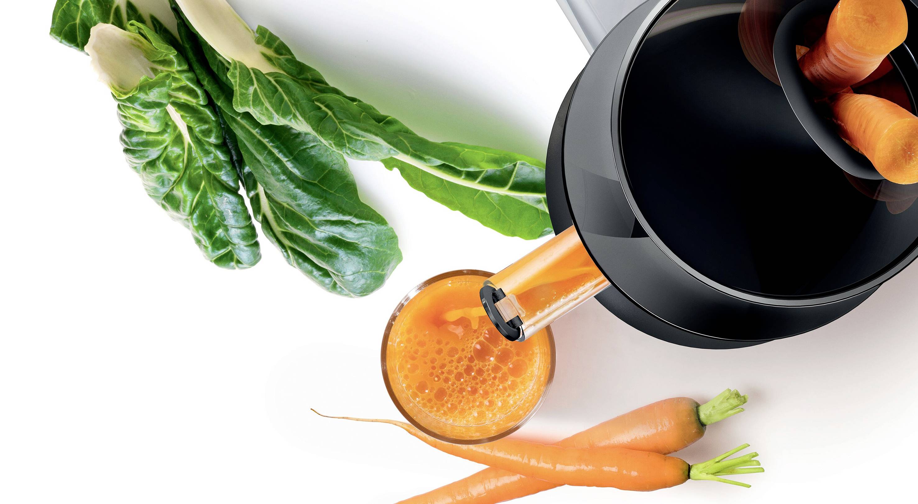 Estrattore con succo di carota appena premuto, accanto carote e verdure a foglia verde.