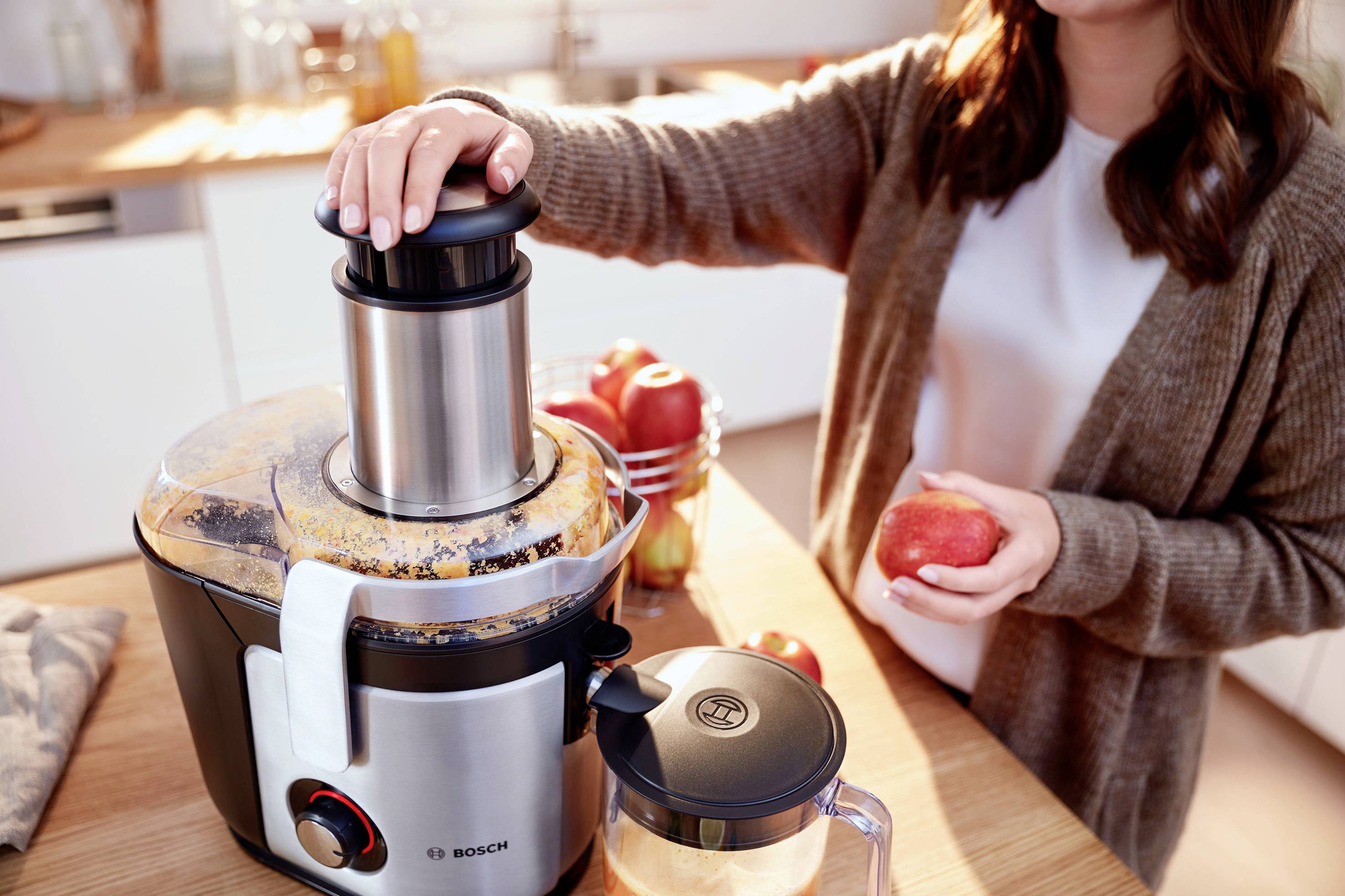 Una donna aziona un estrattore di succo in una cucina e tiene in mano una mela. Sullo sfondo sono visibili alcuni elettrodomestici da cucina.