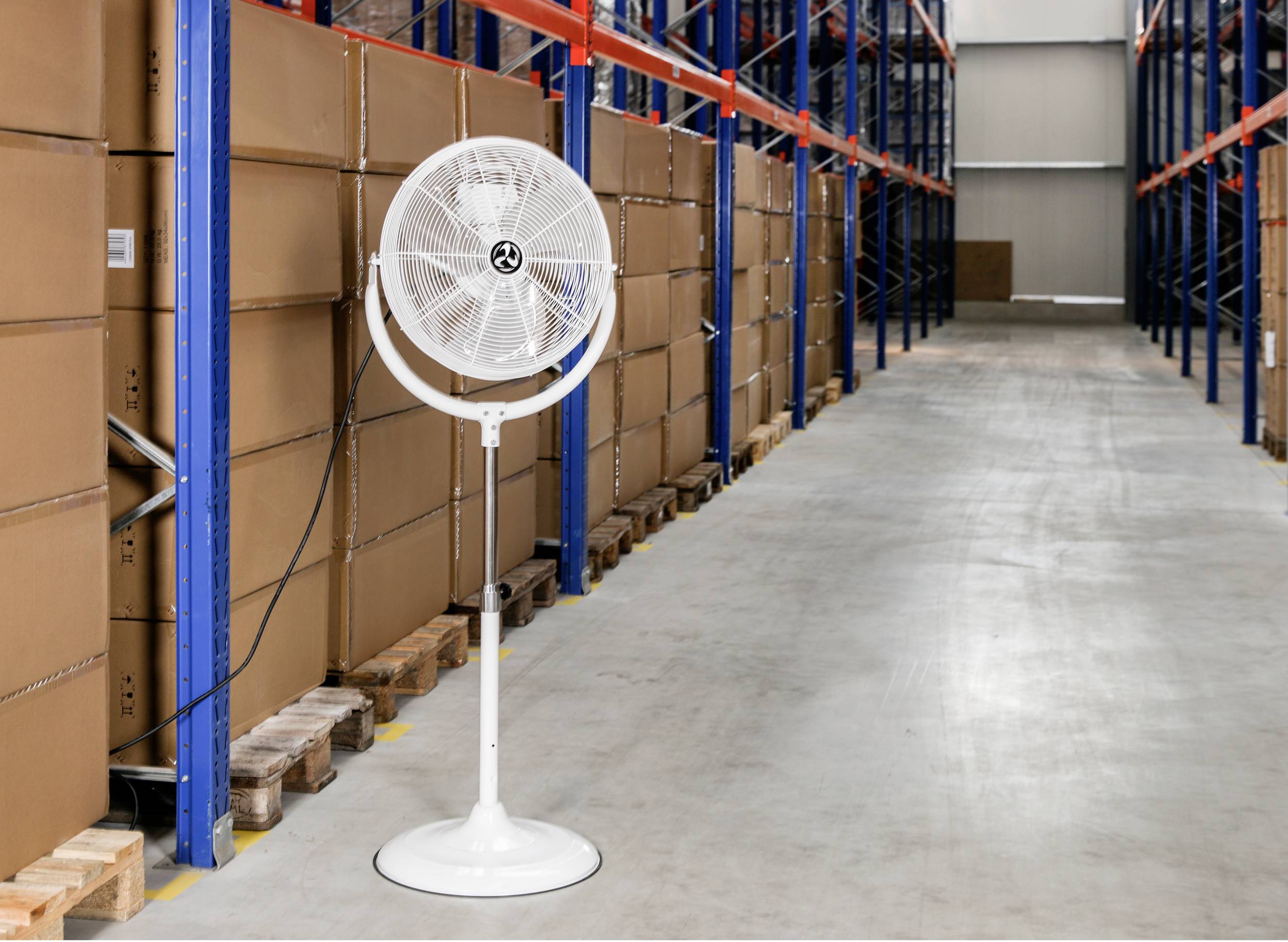 Un ventilatore bianco è posizionato in un grande magazzino accanto a scaffalature alte con scatole marroni. Il pavimento del magazzino è grigio e pulito.