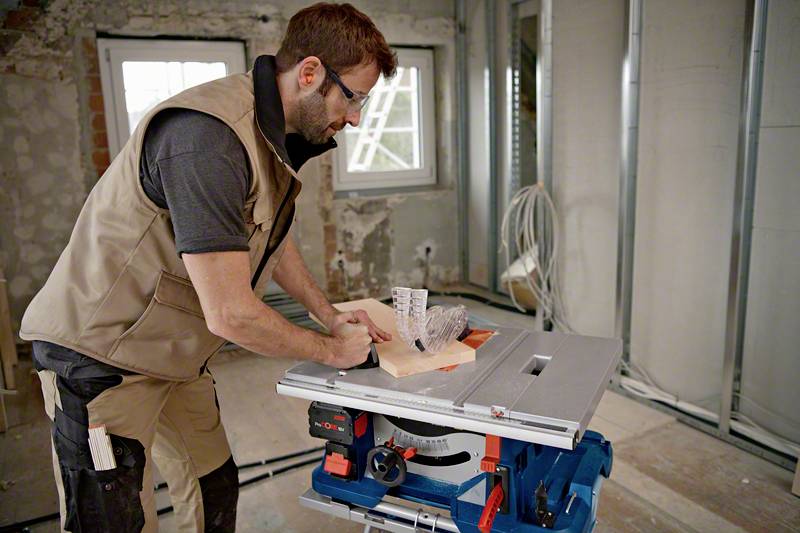 Una persona sta lavorando in un ambiente non finito con una sega da tavolo, tagliando una tavola di legno. Cantiere con cavi esposti e pareti grezze.
