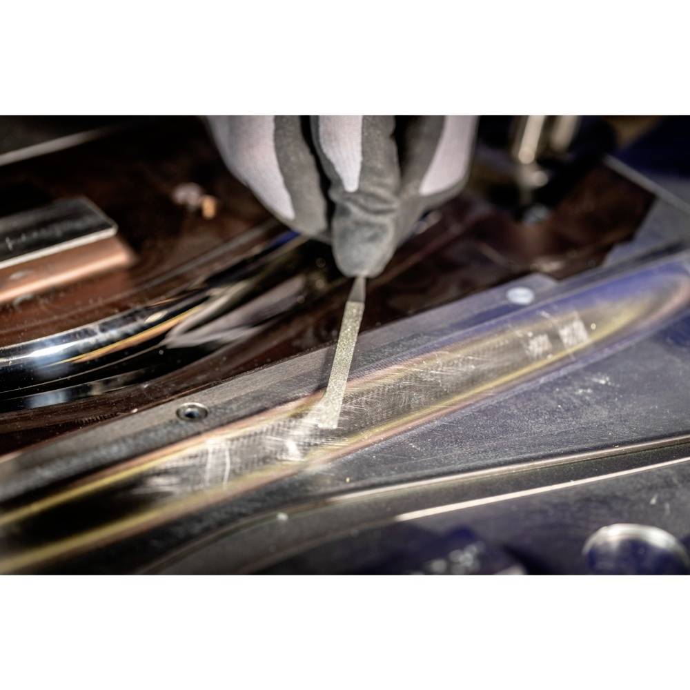 Una mano con un guanto tiene uno strumento per lime che viene utilizzato sul bordo di un oggetto metallico, probabilmente per una lavorazione di precisione.