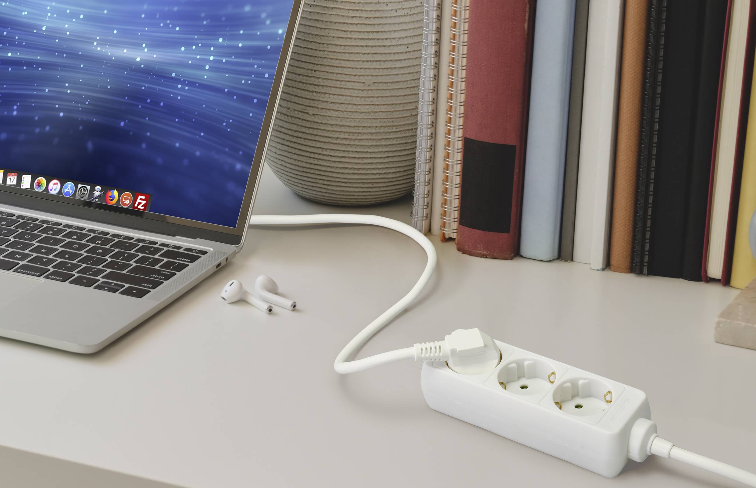 Computer portatile su una scrivania accanto a una multipresa e delle cuffie. Sullo sfondo ci sono dei libri.