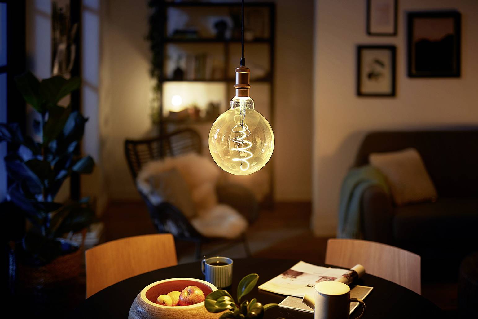 Un ambiente accogliente di un soggiorno con un tavolo rotondo in primo piano, su cui sono posate una ciotola di frutta, tazze e riviste; sullo sfondo uno scaffale illuminato.