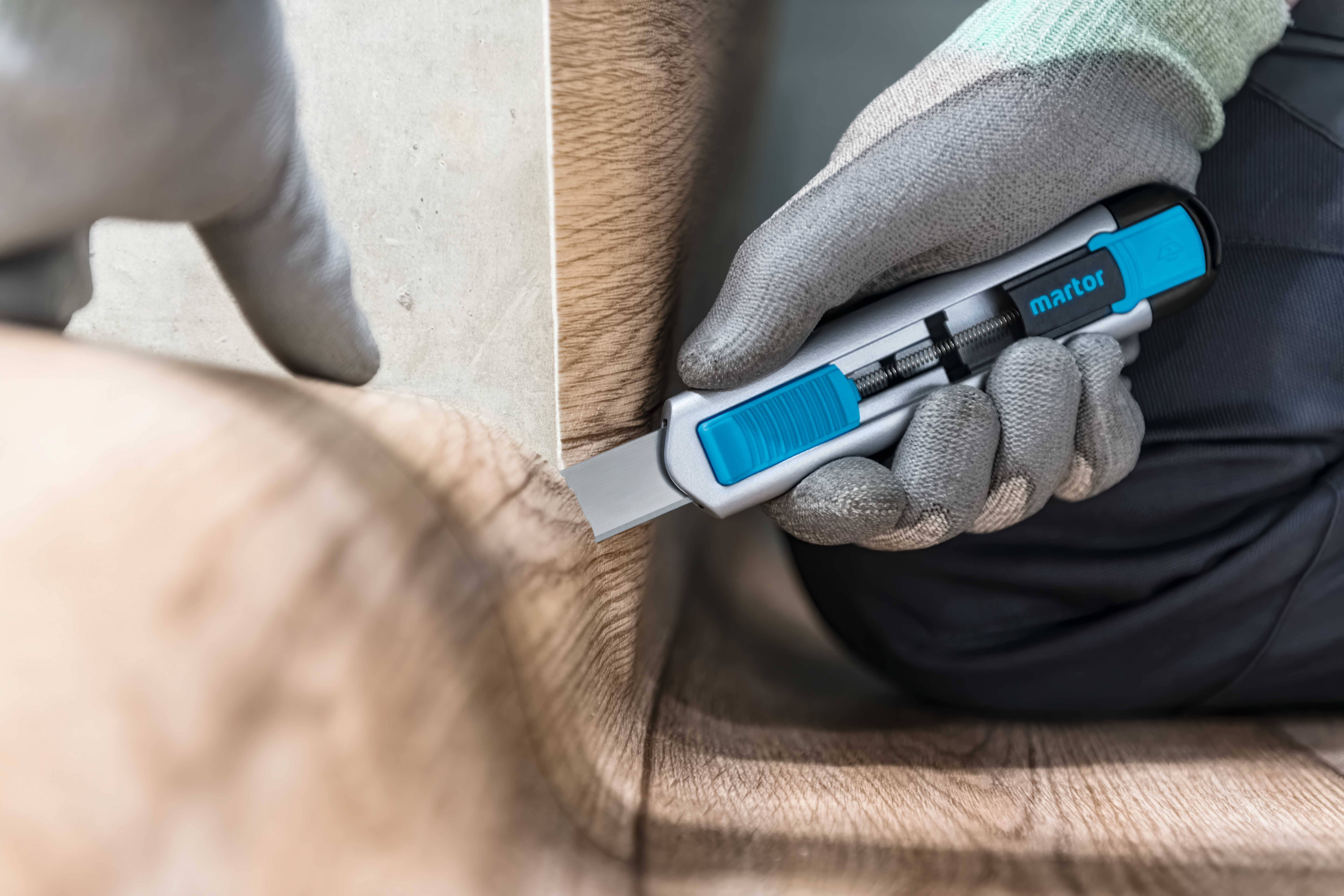 Una persona con i guanti taglia con precisione un rivestimento pavimentale effetto legno con un taglierino blu, per adattarlo agli angoli di una stanza.