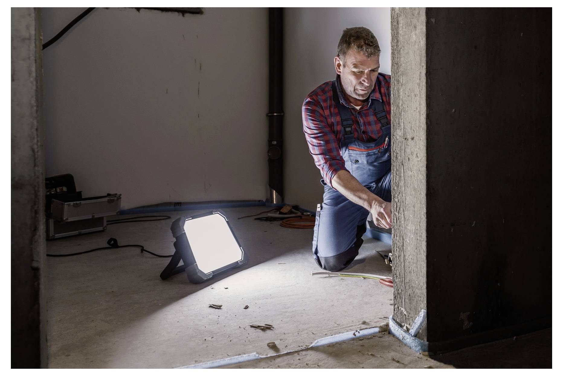 Un uomo si inginocchia accanto a un muro, utilizzando attrezzi per lavorare su cavi elettrici o riparazioni, illuminato da una luminosa lampada da lavoro portatile in una stanza non finita.