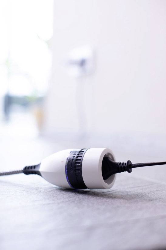 Un connettore bianco e nero è adagiato piatto sul pavimento. Uno sfondo sfocato accenna a una presa a muro.