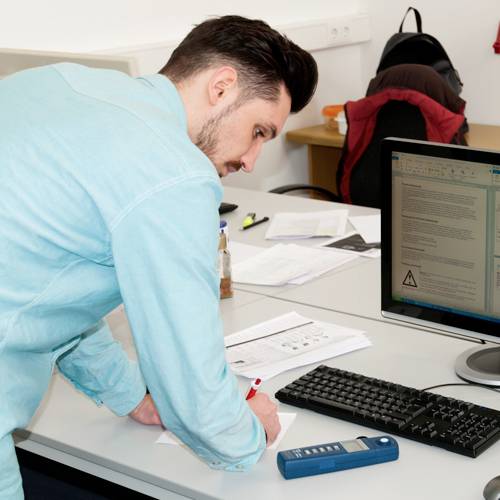 Un uomo con una camicia blu sta contrassegnando un documento su una scrivania con un computer. Atmosfera d'ufficio, con diversi documenti sparsi.