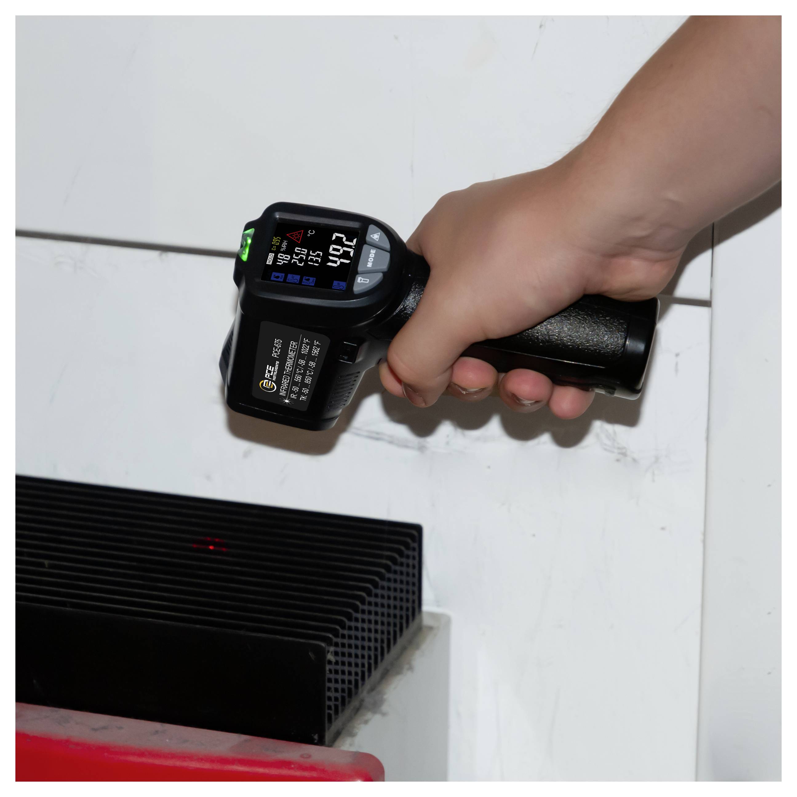 Termometro a infrarossi portatile che misura un dispositivo elettronico nero o una presa di ventilazione su una superficie bianca, mostrando una lettura di temperatura.