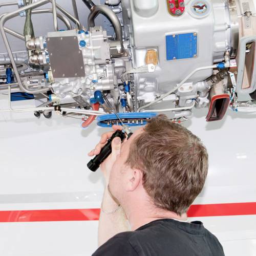Un tecnico ispeziona il componente meccanico complesso di un motore aereo dal basso, utilizzando una torcia elettrica.
