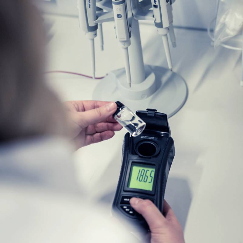 Una persona tiene un piccolo recipiente e misura con un dispositivo digitale che mostra una lettura di 1,8065. Sullo sfondo, attrezzature da laboratorio.