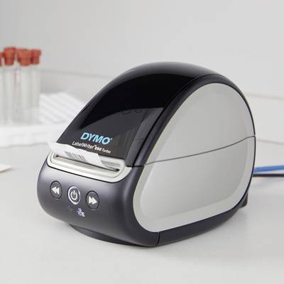 Stampante per etichette sulla scrivania, pronta all'uso. Sullo sfondo sono visibili provette da laboratorio sfocate.