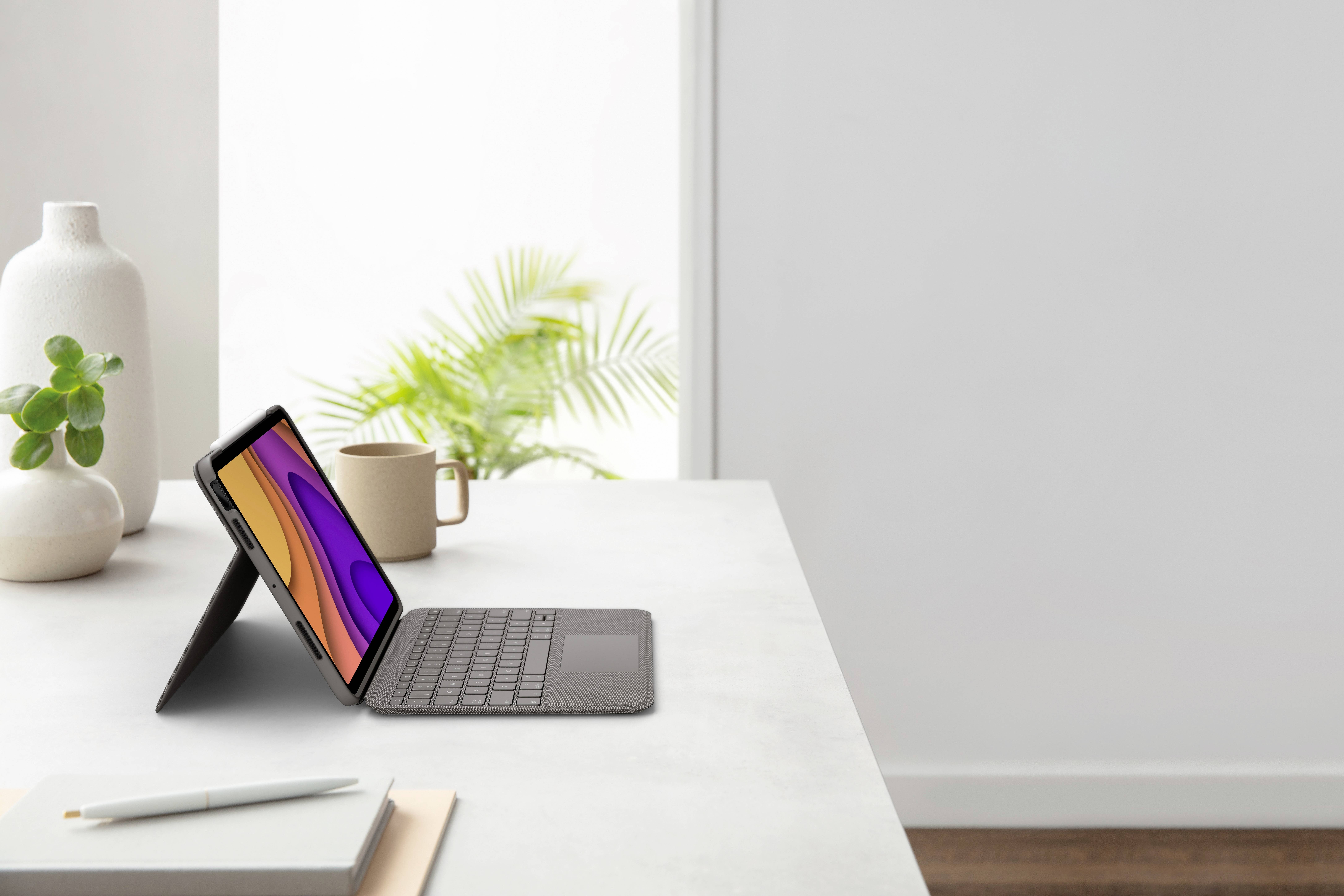 Tablet in custodia-tastiera su un tavolo, accanto a un vaso e una pianta in vaso, davanti a una finestra luminosa. Ambiente di lavoro moderno.