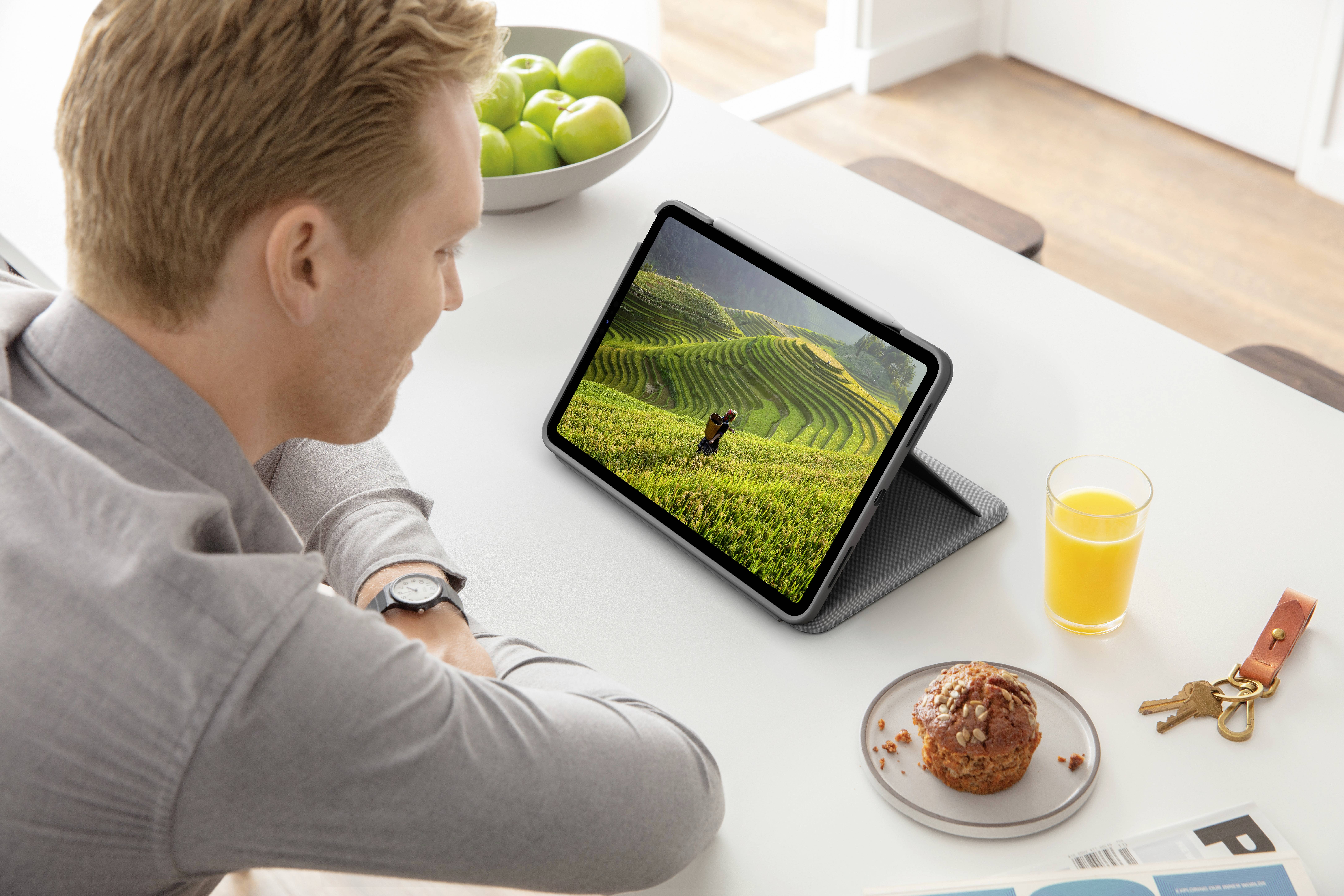 Un uomo guarda un tablet con un'immagine di campi verdi. Sul tavolo ci sono un bicchiere di succo d'arancia, un dolce e un mazzo di chiavi.
