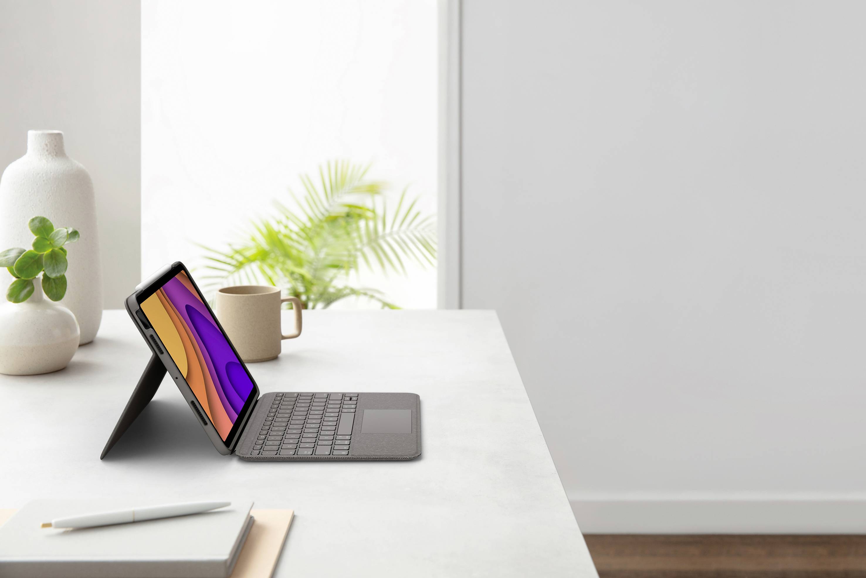 Un tablet con tastiera collegata è posizionato su una scrivania moderna e luminosa, accanto a una tazza, una pianta e una penna.