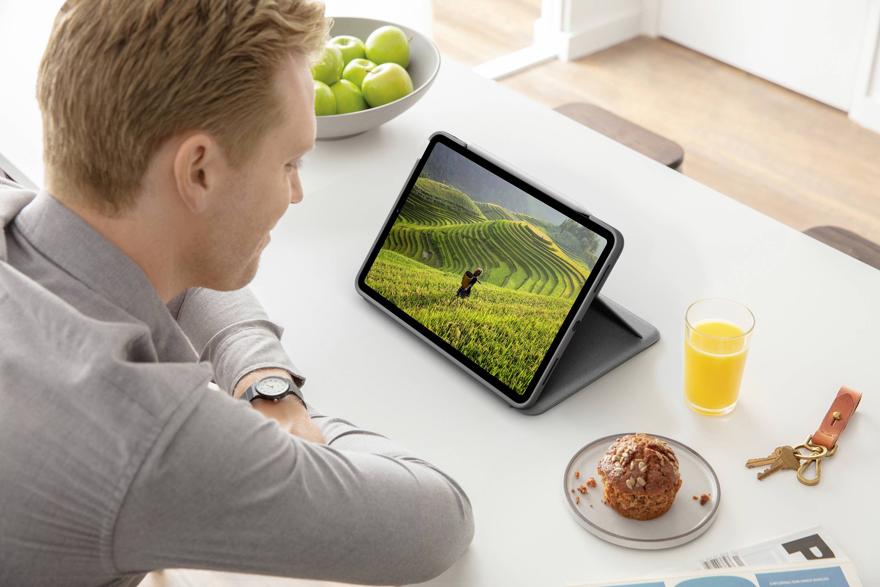 Un uomo seduto al tavolo guarda un tablet che mostra un'immagine di colline verdi. Sul tavolo ci sono un bicchiere di succo e un dolce.