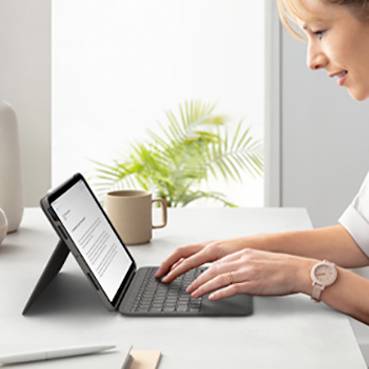 Una donna sta utilizzando un tablet con tastiera su un tavolo. Sta digitando. Sullo sfondo, una pianta e una tazza.