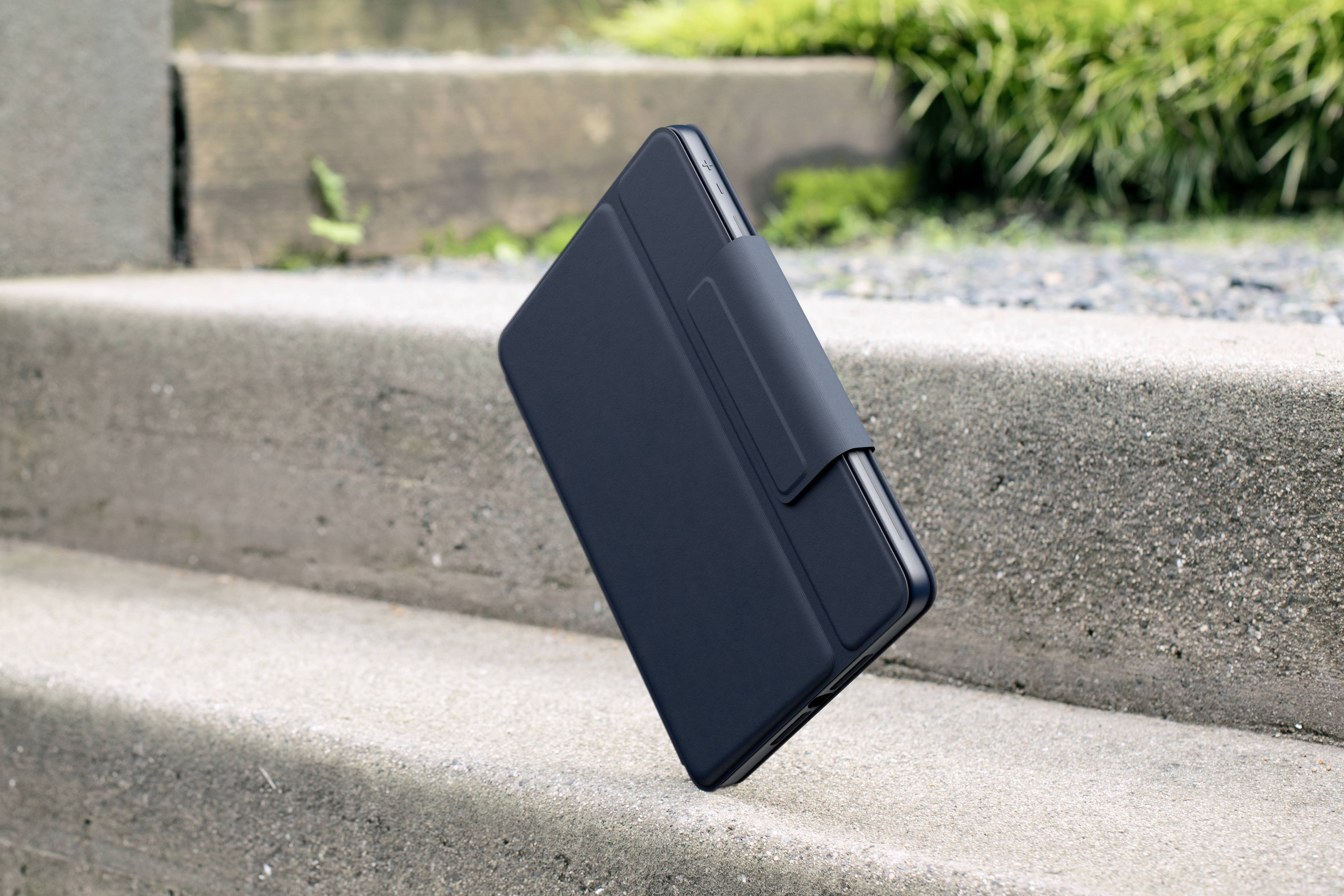 Un tablet chiuso in un involucro nero aleggia sopra una scalinata di pietra all'aperto. Sullo sfondo sono visibili piante verdi.