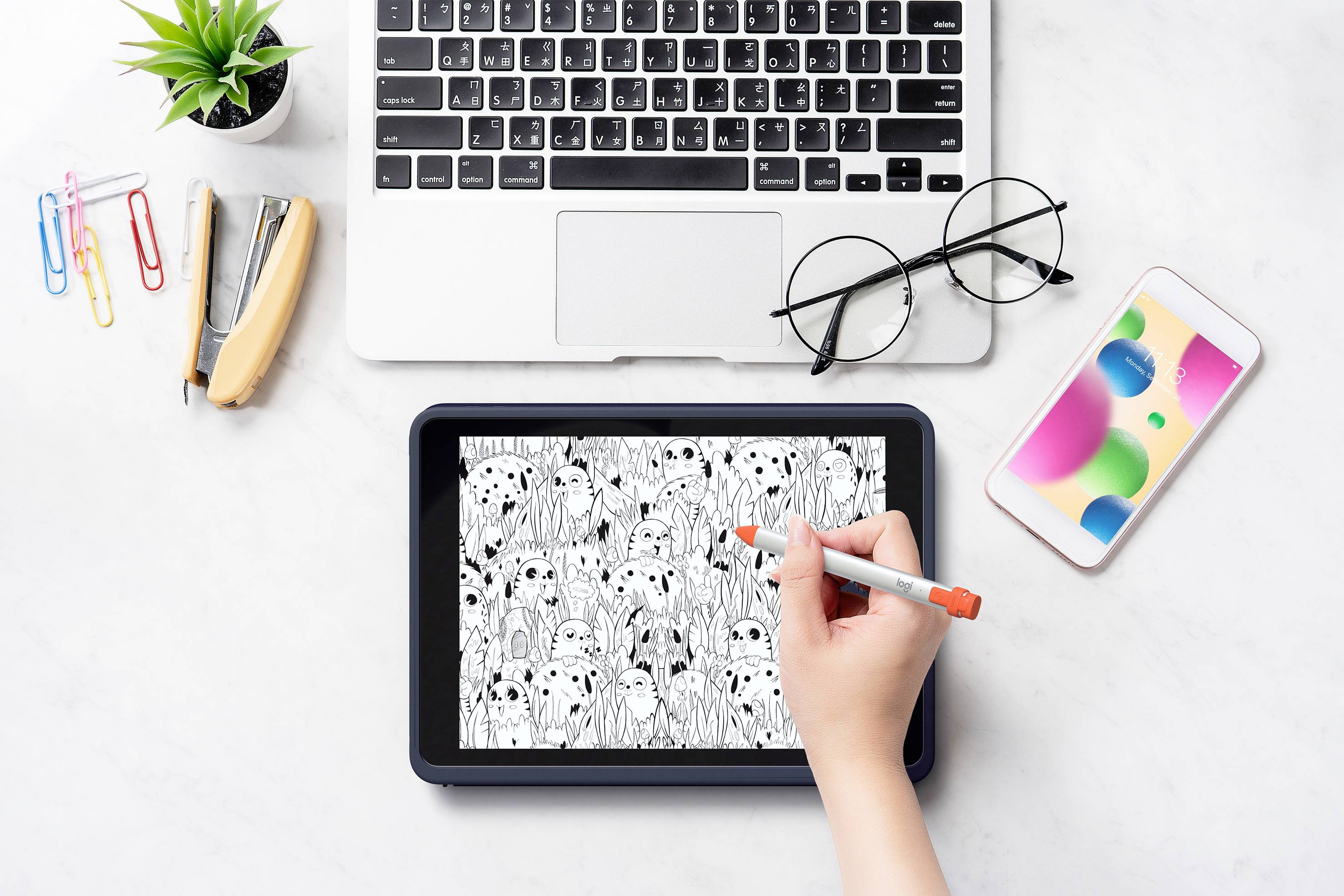 Una mano disegna un motivo in bianco e nero su un tablet. Sullo sfondo sono visibili un laptop, un paio di occhiali e uno smartphone.