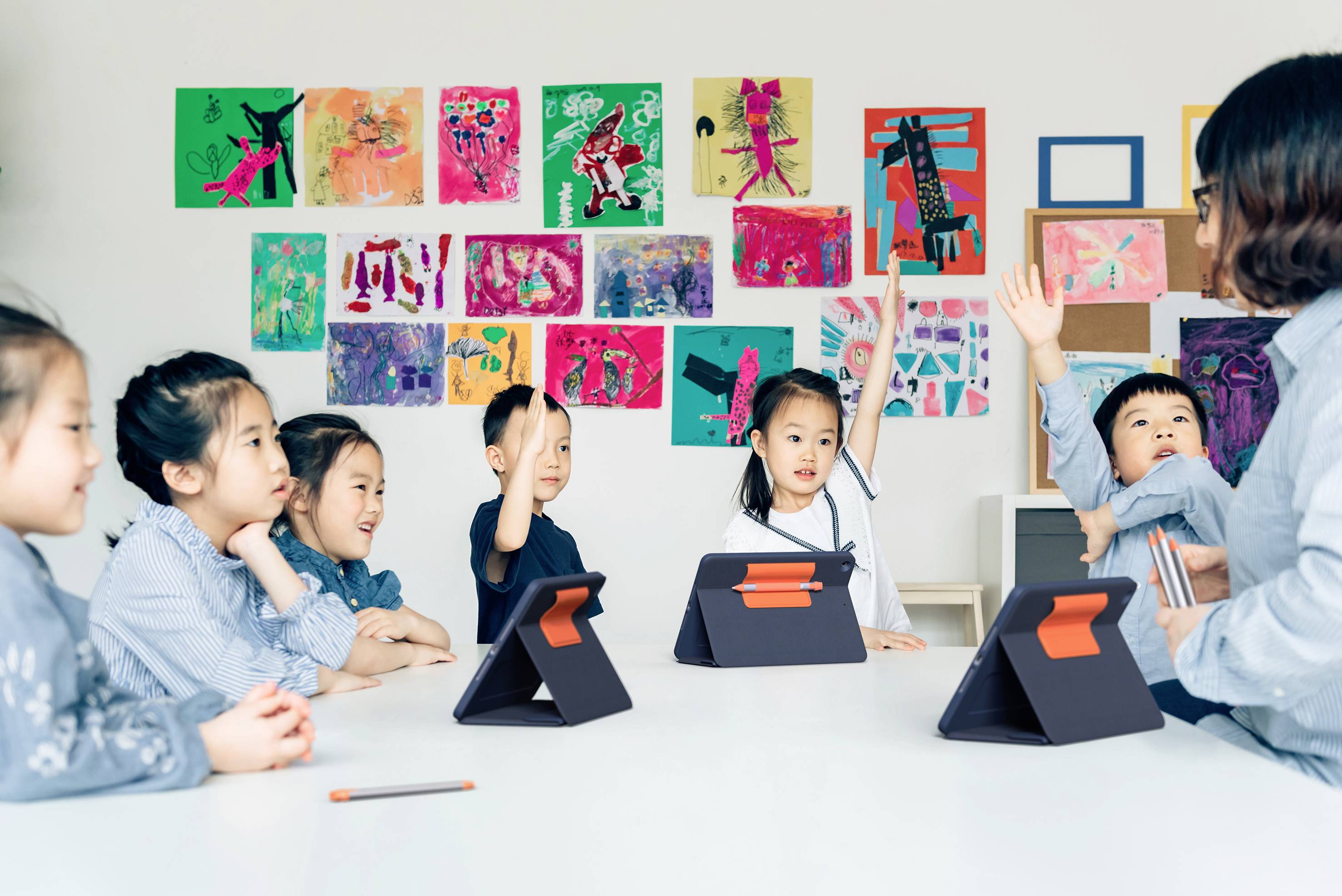Un'insegnante sta istruendo un gruppo di bambini seduti a un tavolo con dei tablet. Alcuni bambini alzano la mano per intervenire. Sullo sfondo, disegni colorati sono appesi al muro.