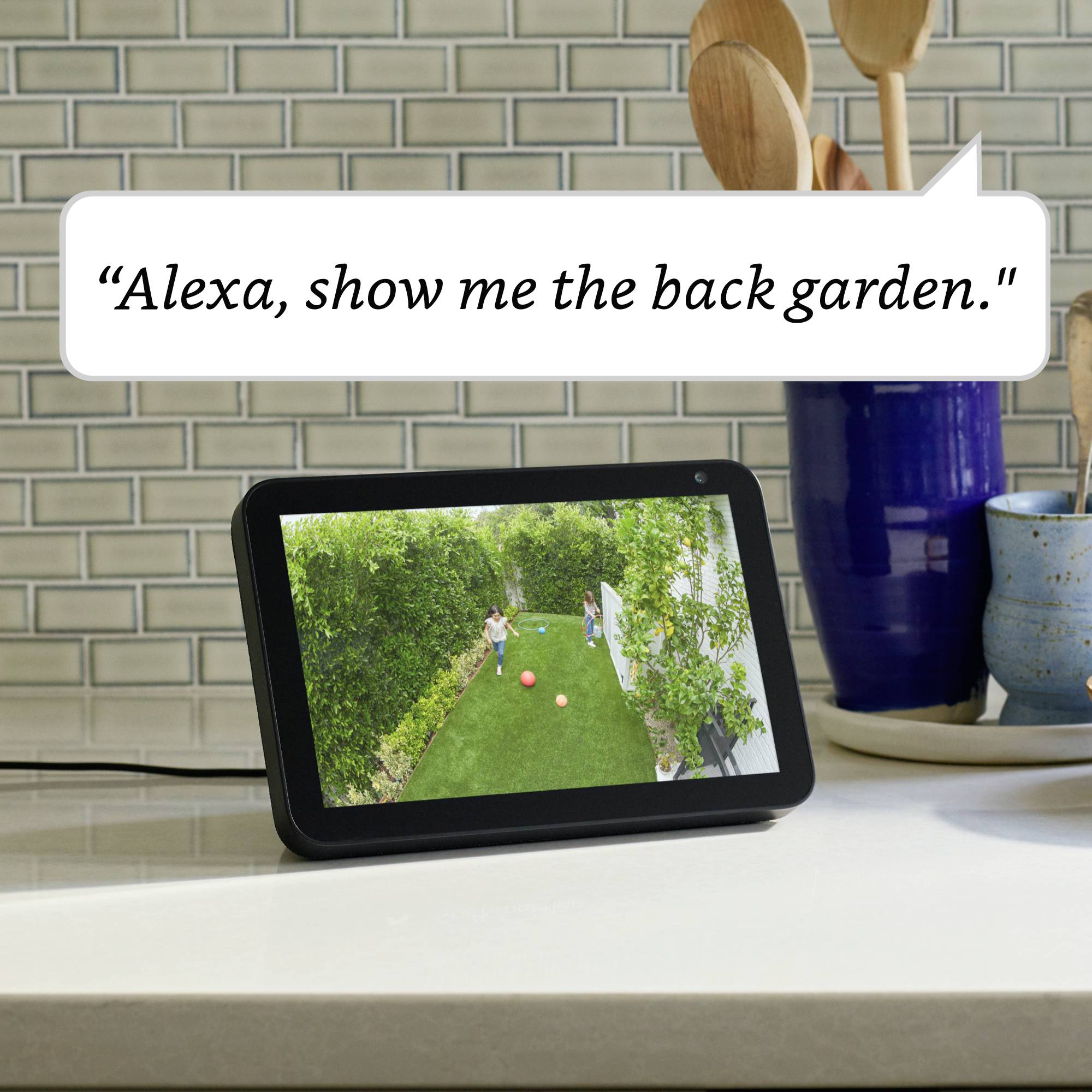 Un display intelligente mostra un'immagine del giardino. Sullo sfondo c'è un vaso blu. Il testo 'Alexa, mostrami il giardino posteriore.' è visibile.