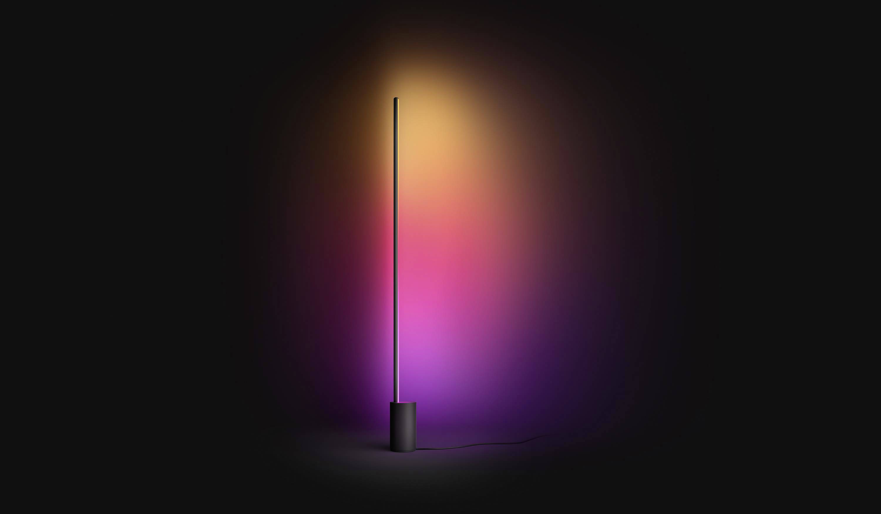 Lampada da terra con un lungo e sottile stelo che irradia luce multicolore in giallo, arancione e viola in un ambiente buio. Design moderno e minimalista.