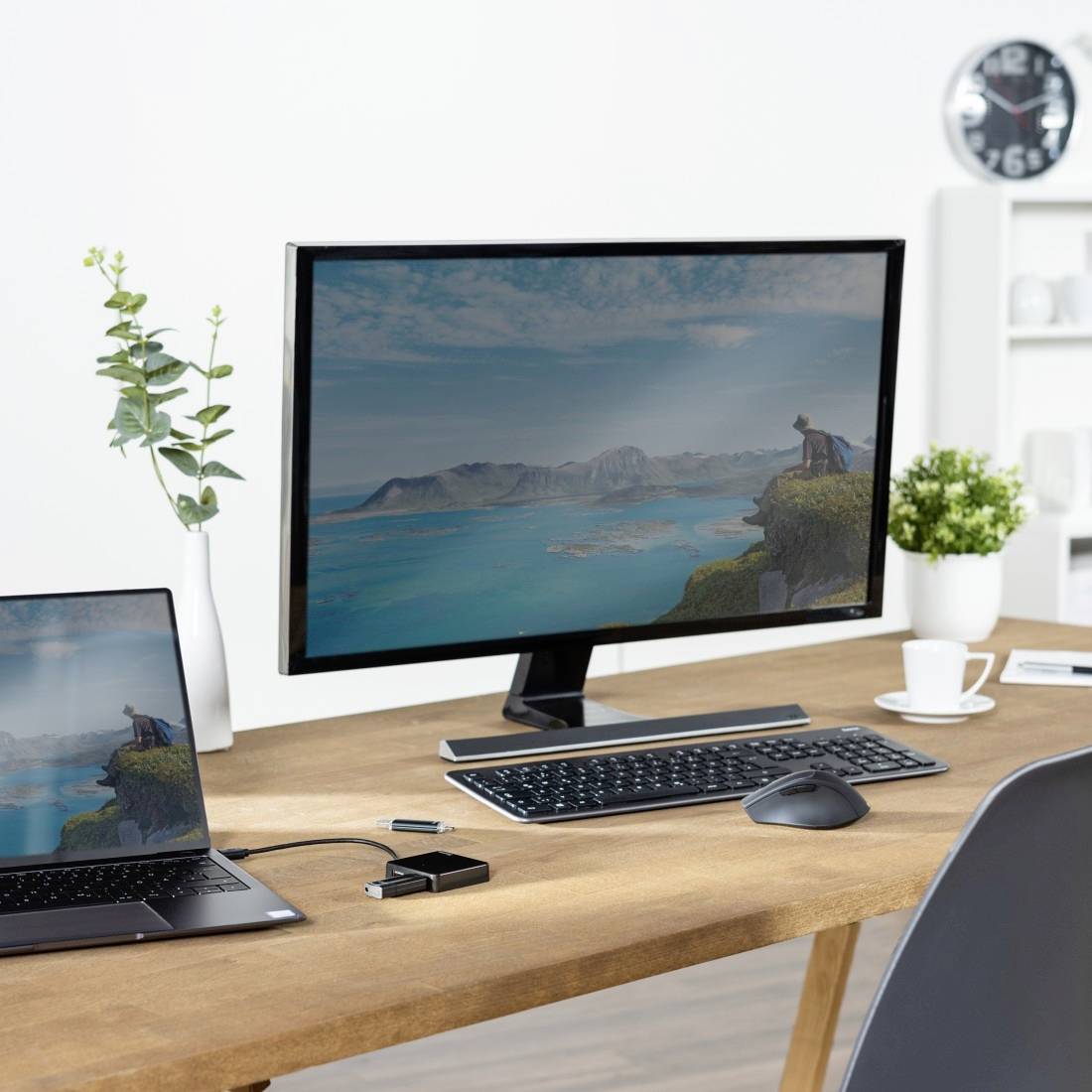 Una scrivania con un laptop e un monitor che mostra un paesaggio. Accanto sono posizionate una tastiera e un mouse. Sullo sfondo si vedono un orologio e degli scaffali.