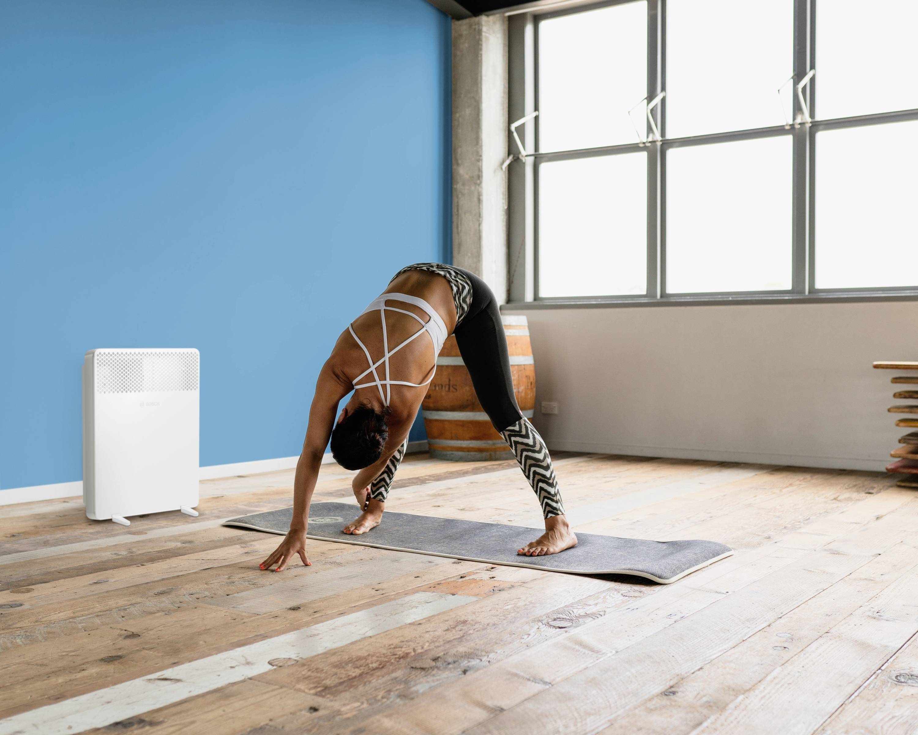 Una persona fa yoga in una stanza con pavimento in legno, ampia vetrata e un accento blu sulla parete; un deumidificatore è posizionato a sinistra.