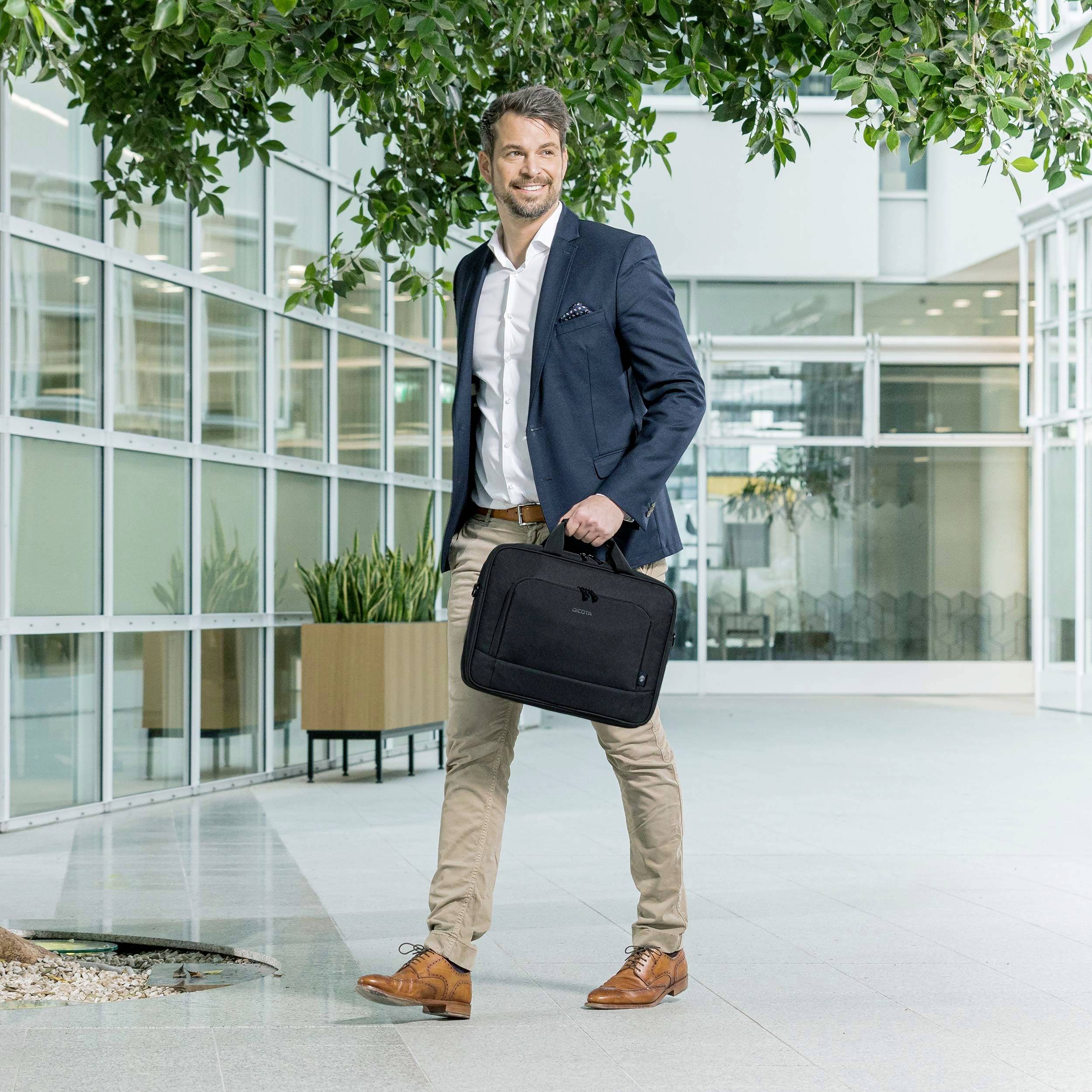 Uomo in abbigliamento da lavoro sorride mentre attraversa un edificio per uffici moderno, portando in mano una borsa nera.