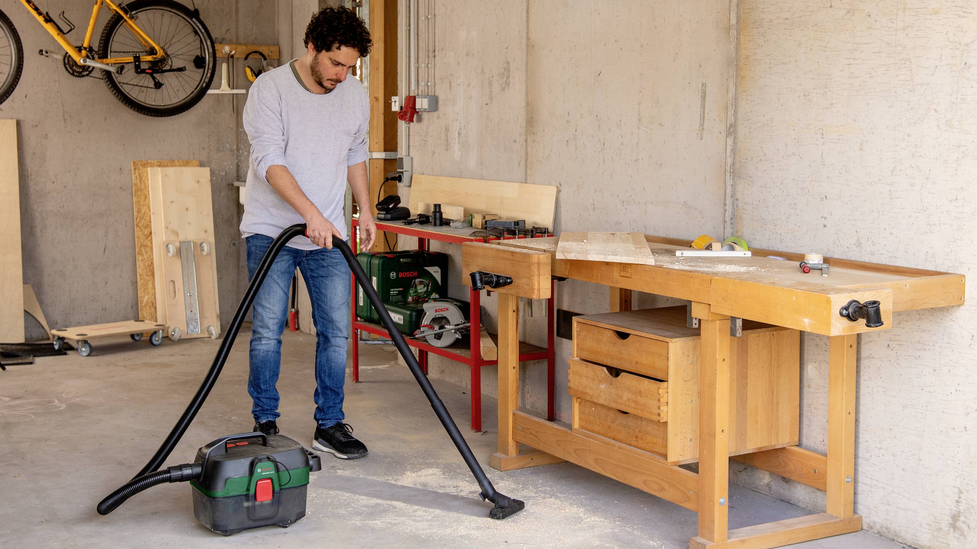 Una persona aspira trucioli di legno in un laboratorio con un aspiratore industriale accanto a un banco da lavoro.
