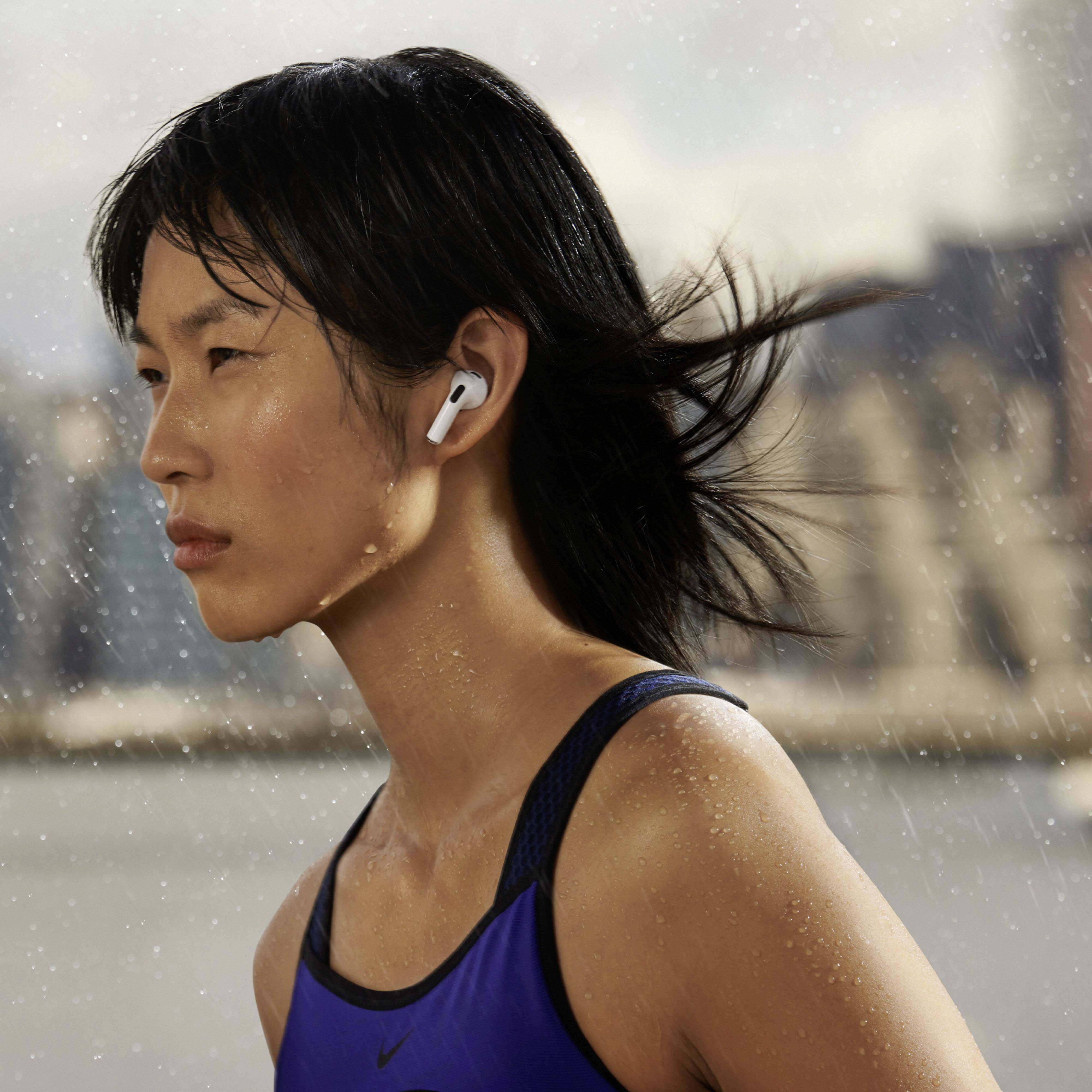 Una donna fa jogging sotto una pioggia leggera, indossando cuffie wireless. Guarda dritta davanti a sé con concentrazione. Sullo sfondo, gli edifici appaiono sfocati.