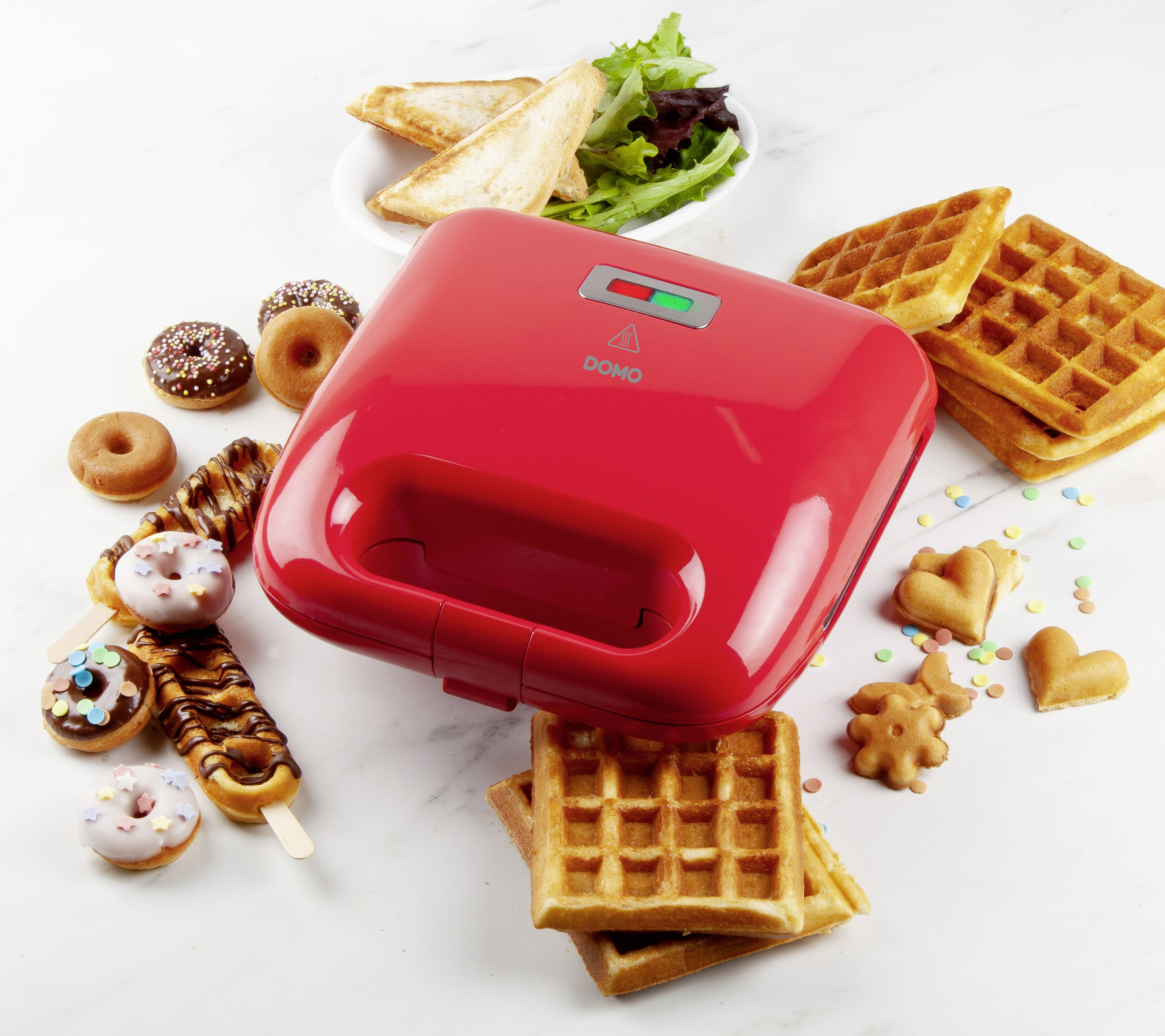Macchina per sandwich rossa accanto a waffle, biscotti e mini ciambelle su un tavolo. Sullo sfondo un piatto con sandwich e insalata.
