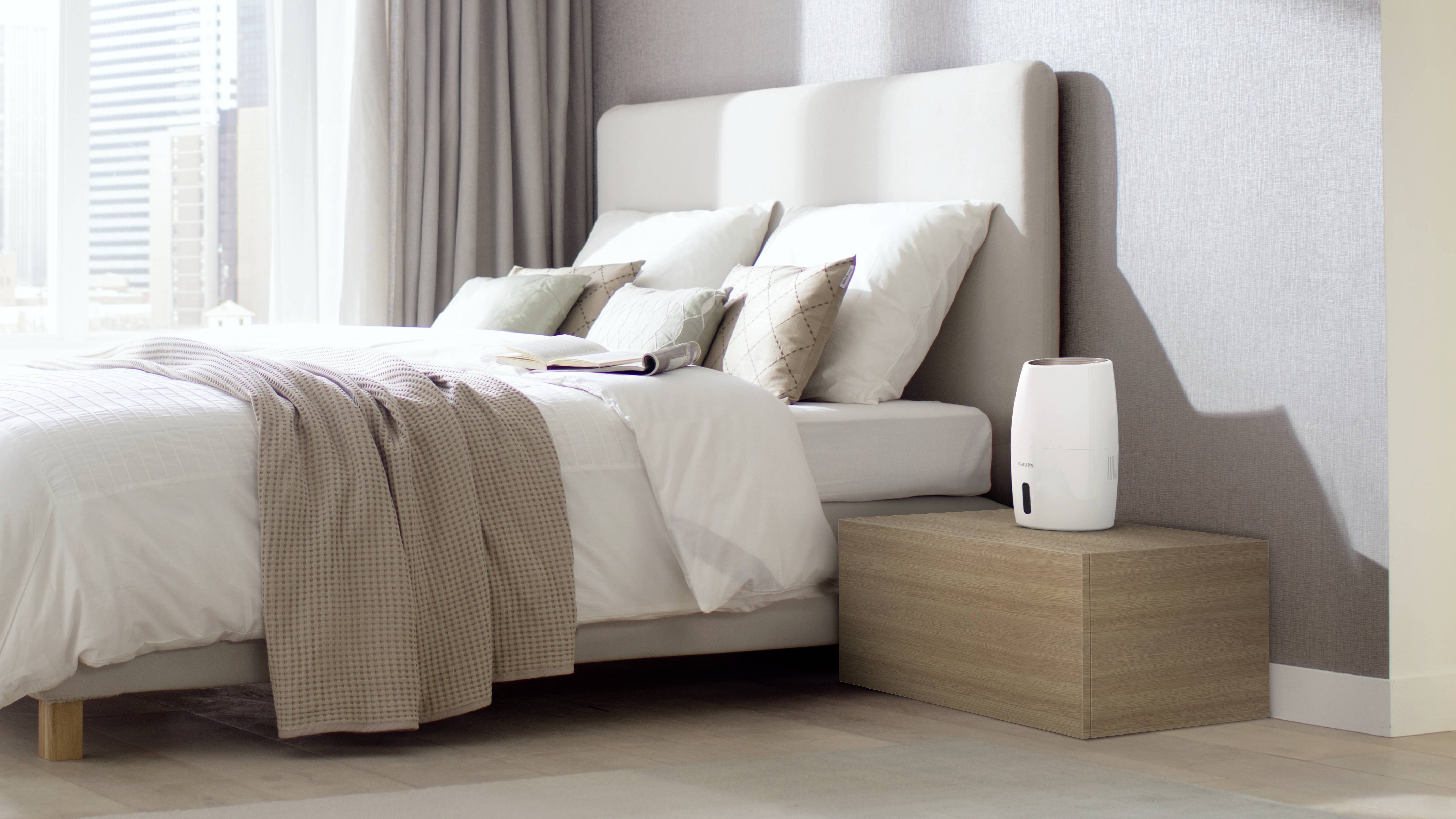 Camera da letto con letto matrimoniale, molti cuscini, piumone e comodino con umidificatore bianco, accanto a finestre alte con tende.