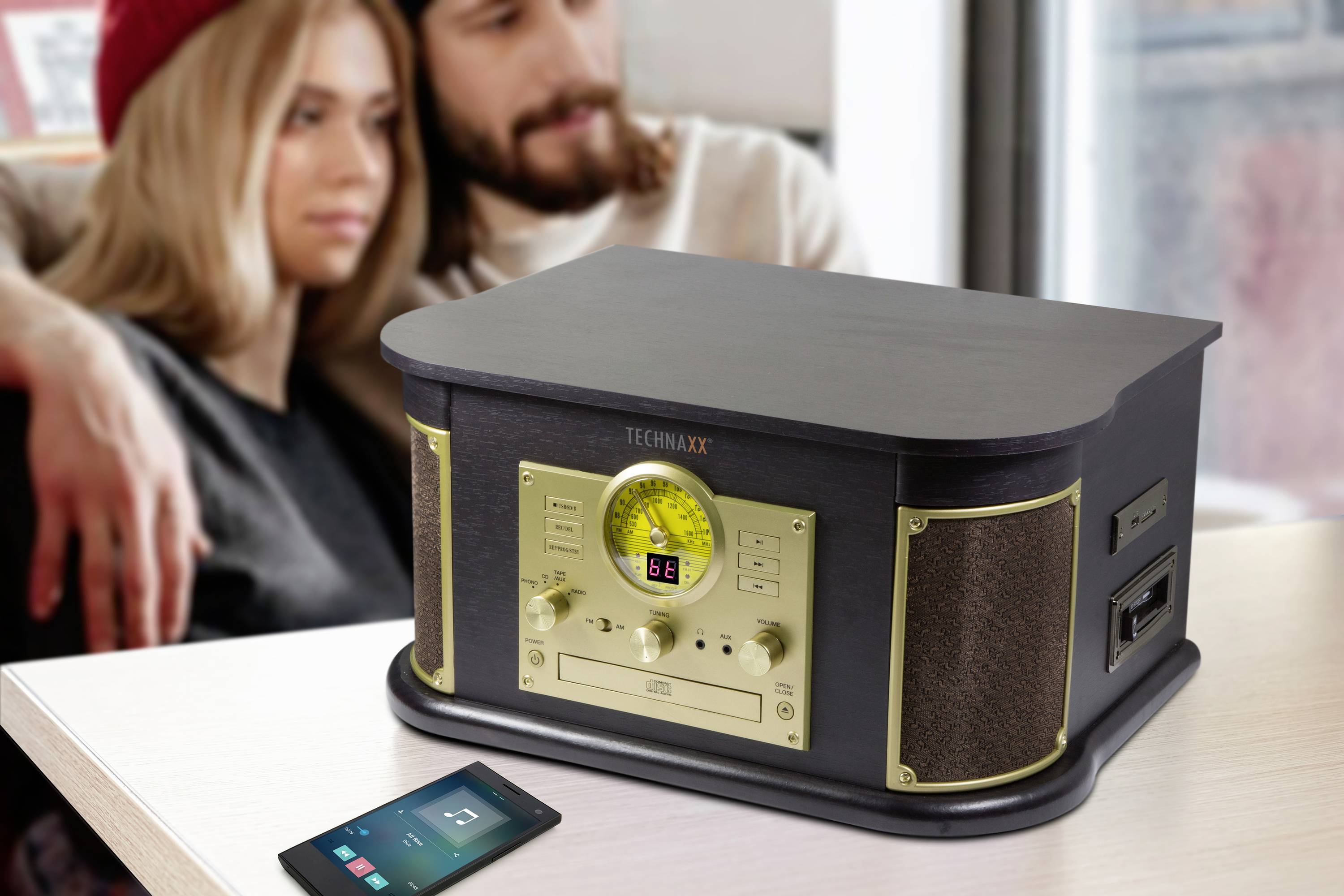 Un lettore CD retrò con radio e altoparlanti è posizionato su un tavolo. Sullo sfondo, una coppia è seduta rilassata fianco a fianco.