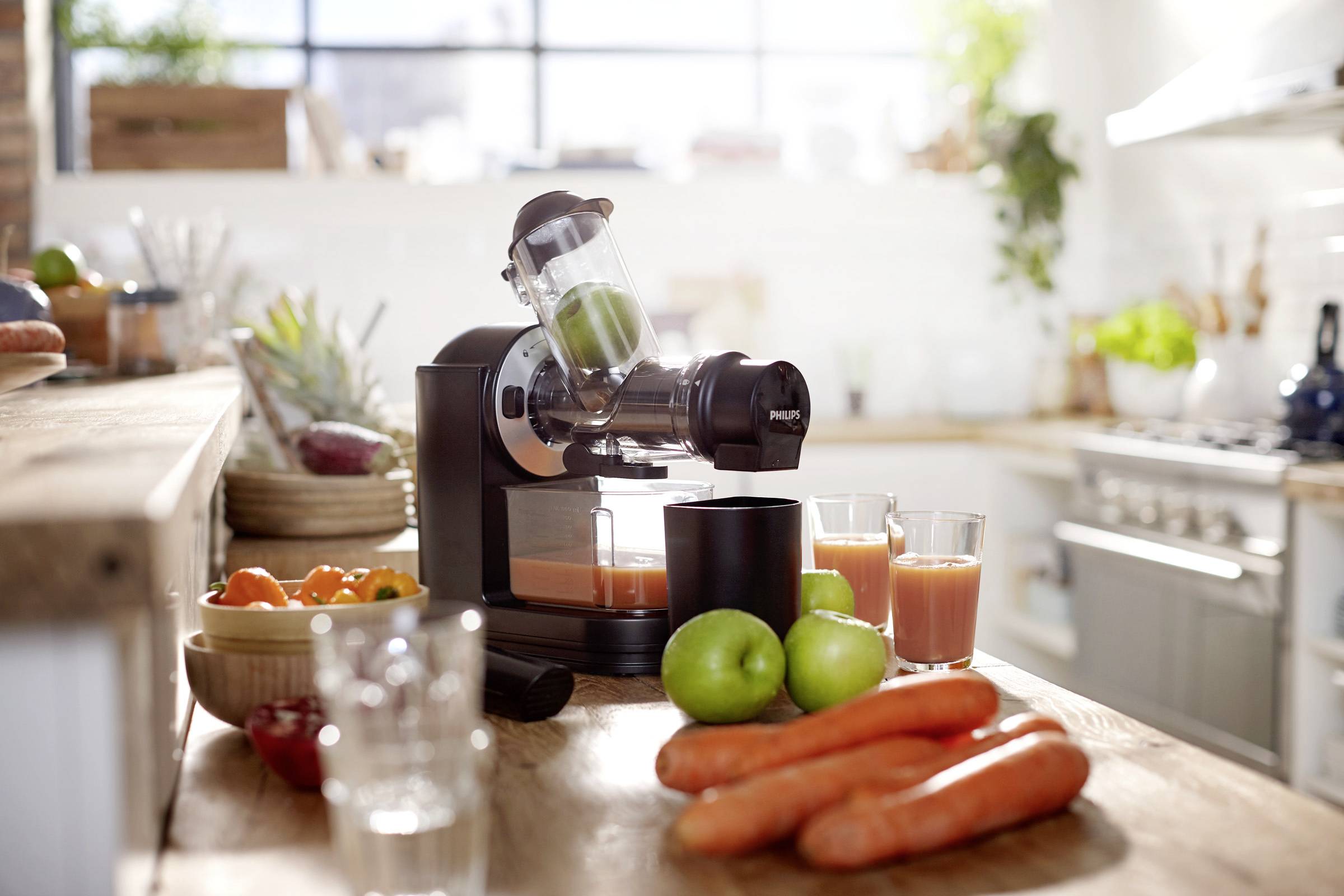Estrattore di succo sul bancone della cucina con succo di carote fresco e verdure. Cucina luminosa sullo sfondo, con frutta e verdura disposte in modo decorativo.