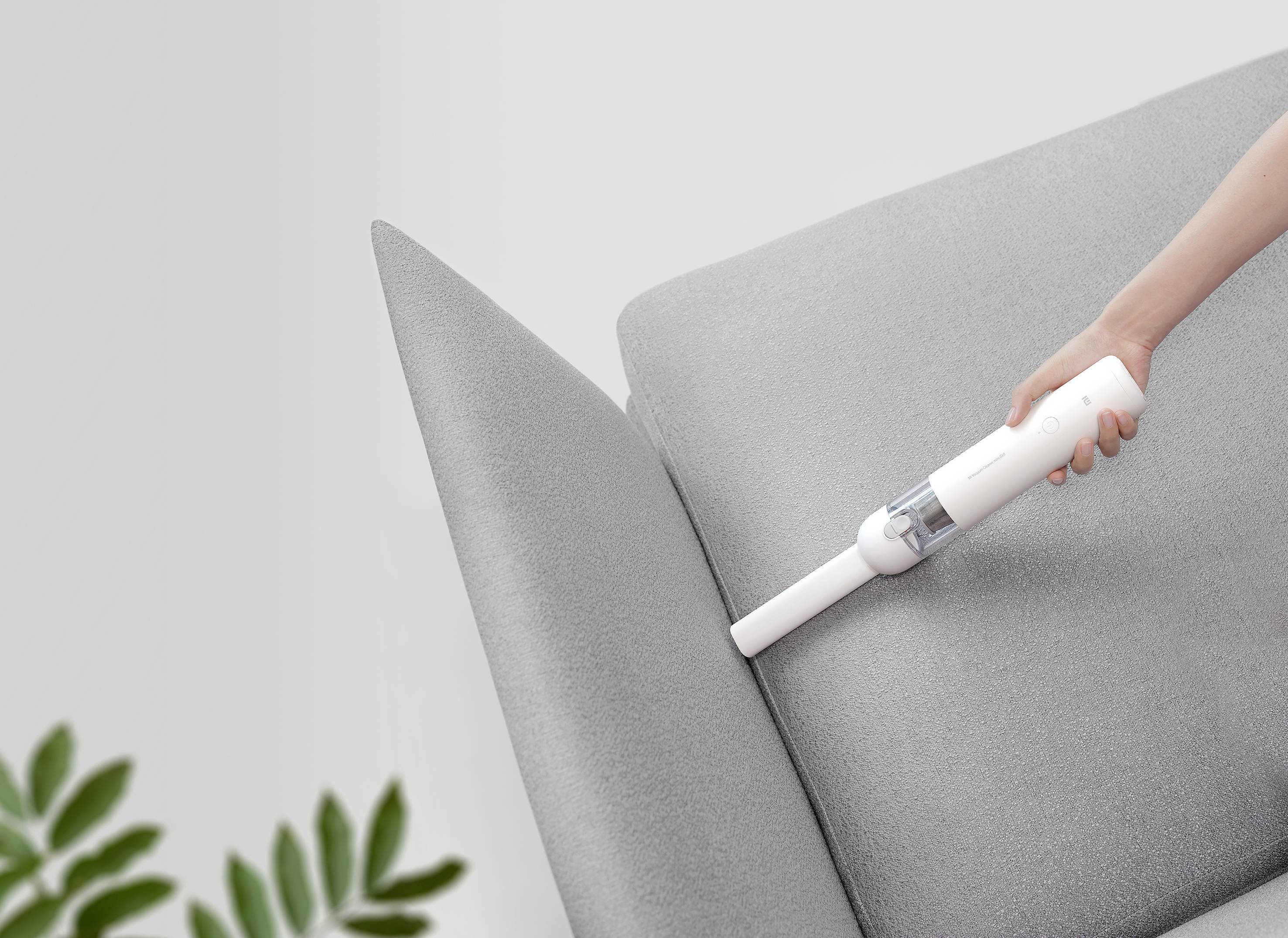 Una persona pulisce una fessura del divano con un piccolo aspirapolvere manuale bianco. In basso a sinistra dell'immagine sono visibili delle foglie di pianta sfocate.