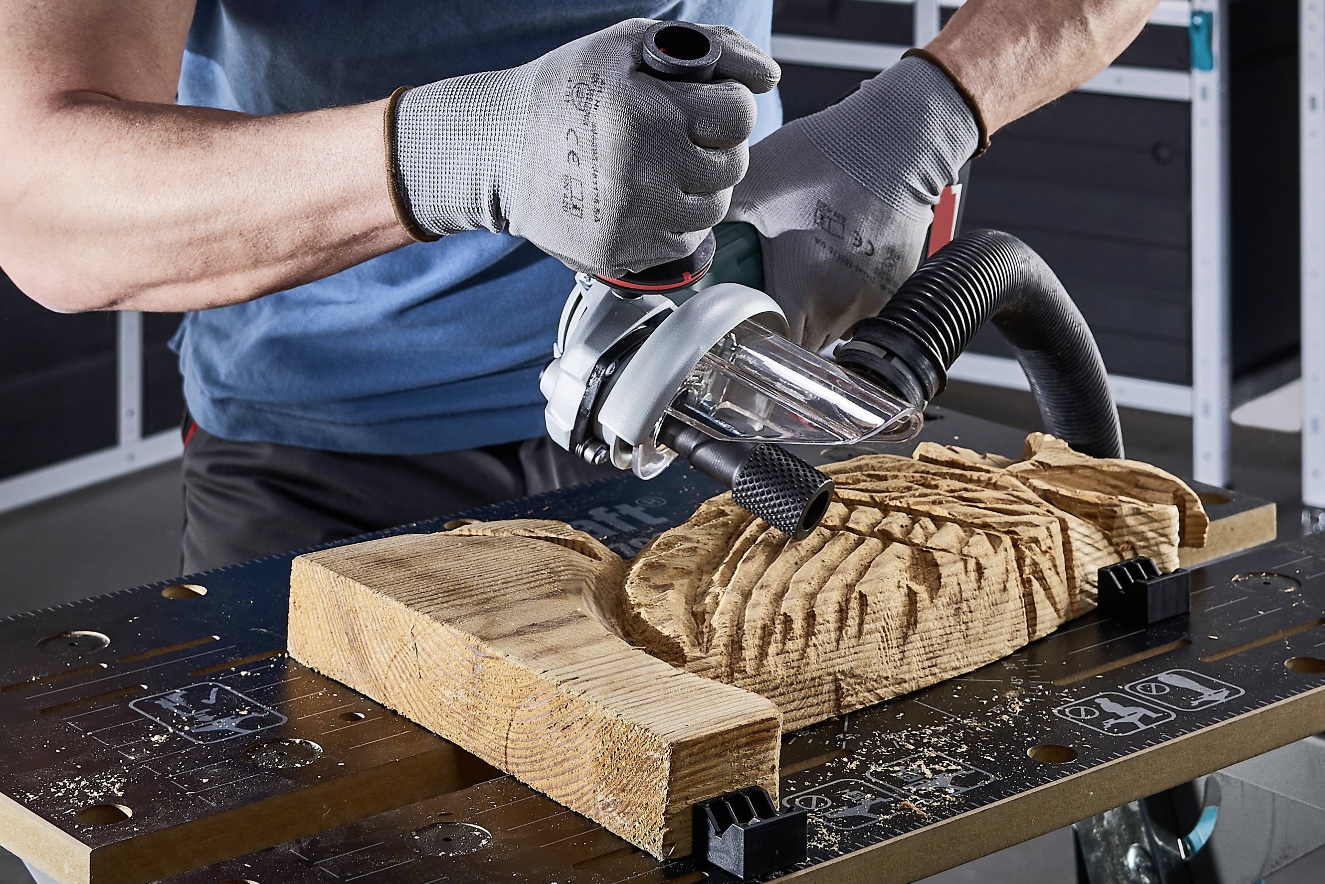Un uomo sta lavorando un pezzo di legno con una fresa elettrica. Indossa guanti da lavoro. Trucioli di legno sono visibili.