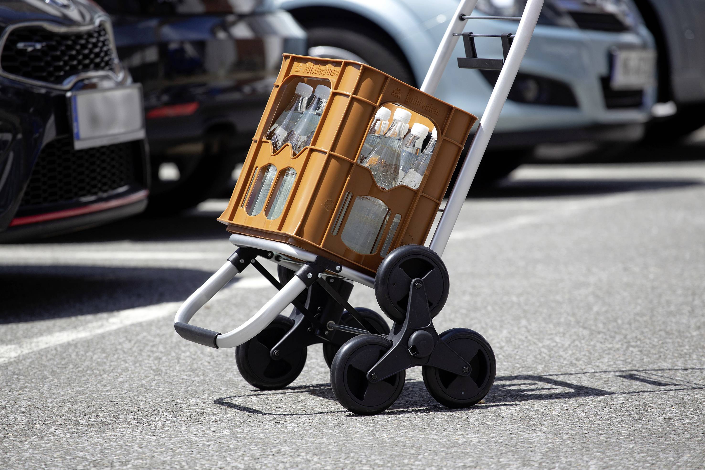Un carretto a mano con ruote grandi trasporta una cassa di bevande arancioni in un parcheggio, con auto sullo sfondo.