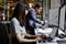 Due persone lavorano a postazioni informatiche. In primo piano una donna con un laptop, sullo sfondo un uomo con un tablet, circondato da monitor.