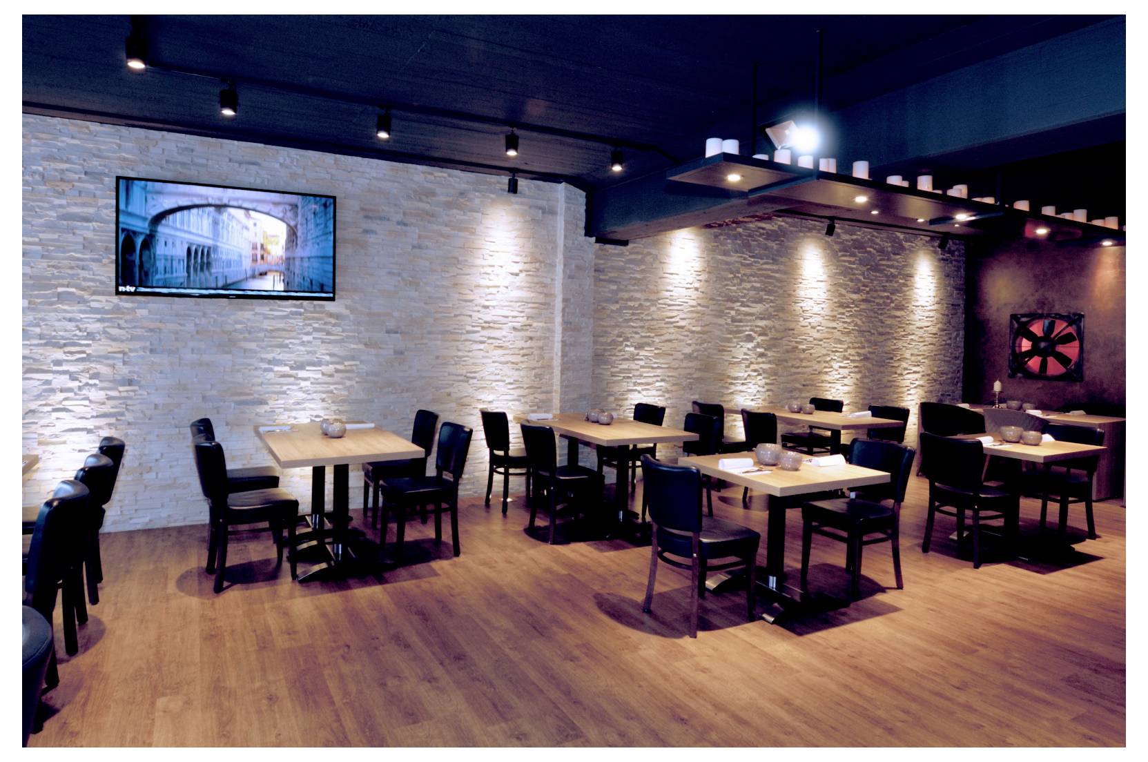 Sala da pranzo moderna e vuota, con tavoli di legno e sedie nere sotto illuminazione a binario, con uno schermo a parete e pareti di pietra testurizzata.