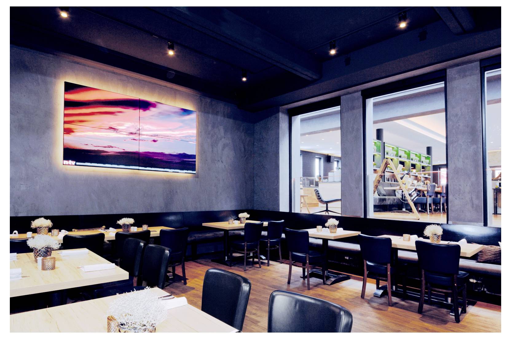 Zona ristorante moderna con sedie nere e tavoli bianchi, una parete con uno schermo che mostra un tramonto e una vista sulla cucina attraverso grandi finestre.