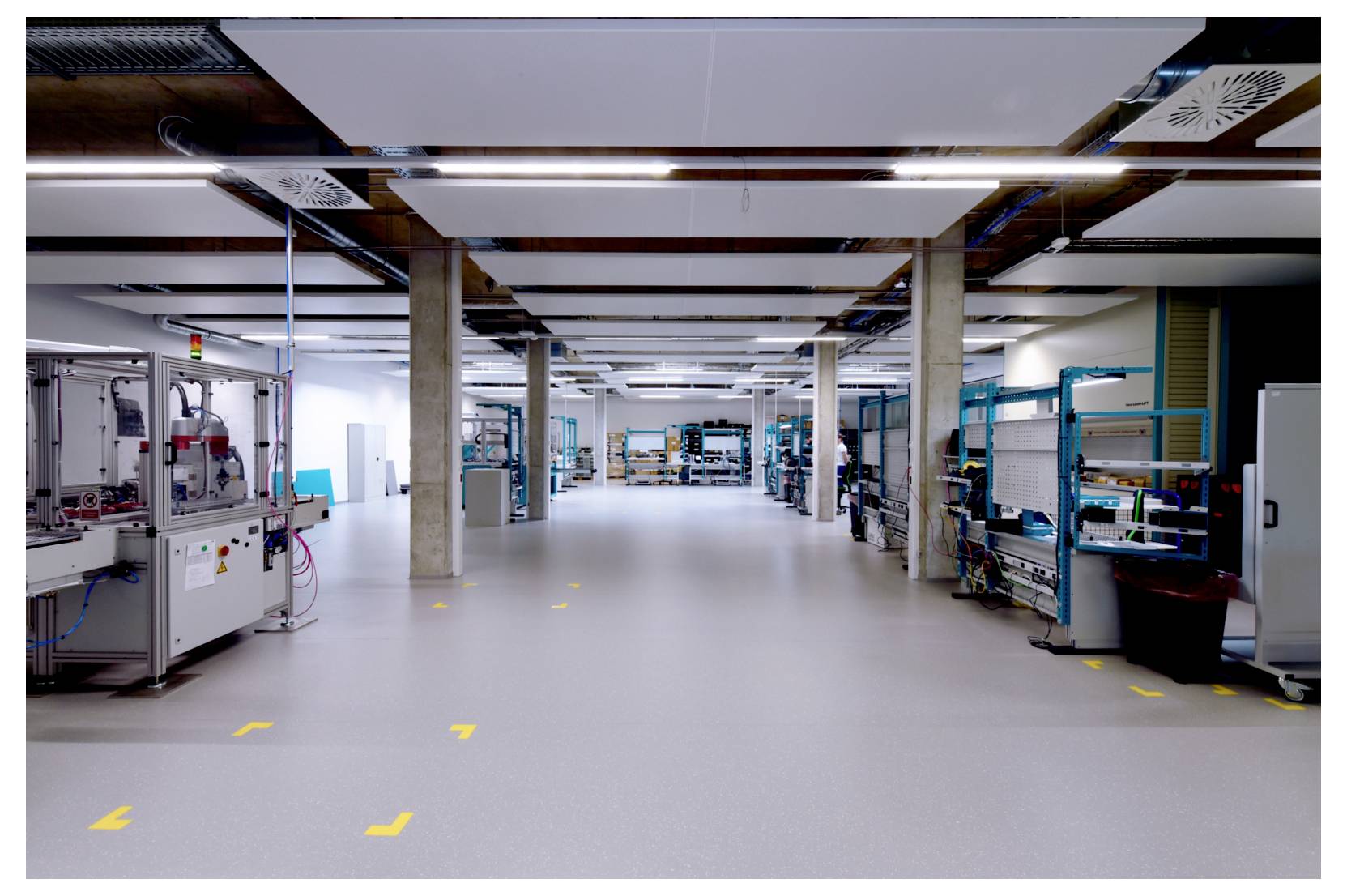Ampio spazio di lavoro industriale con file di macchinari e scaffalature su entrambi i lati, corsia centrale libera e illuminazione intensa dall'alto.