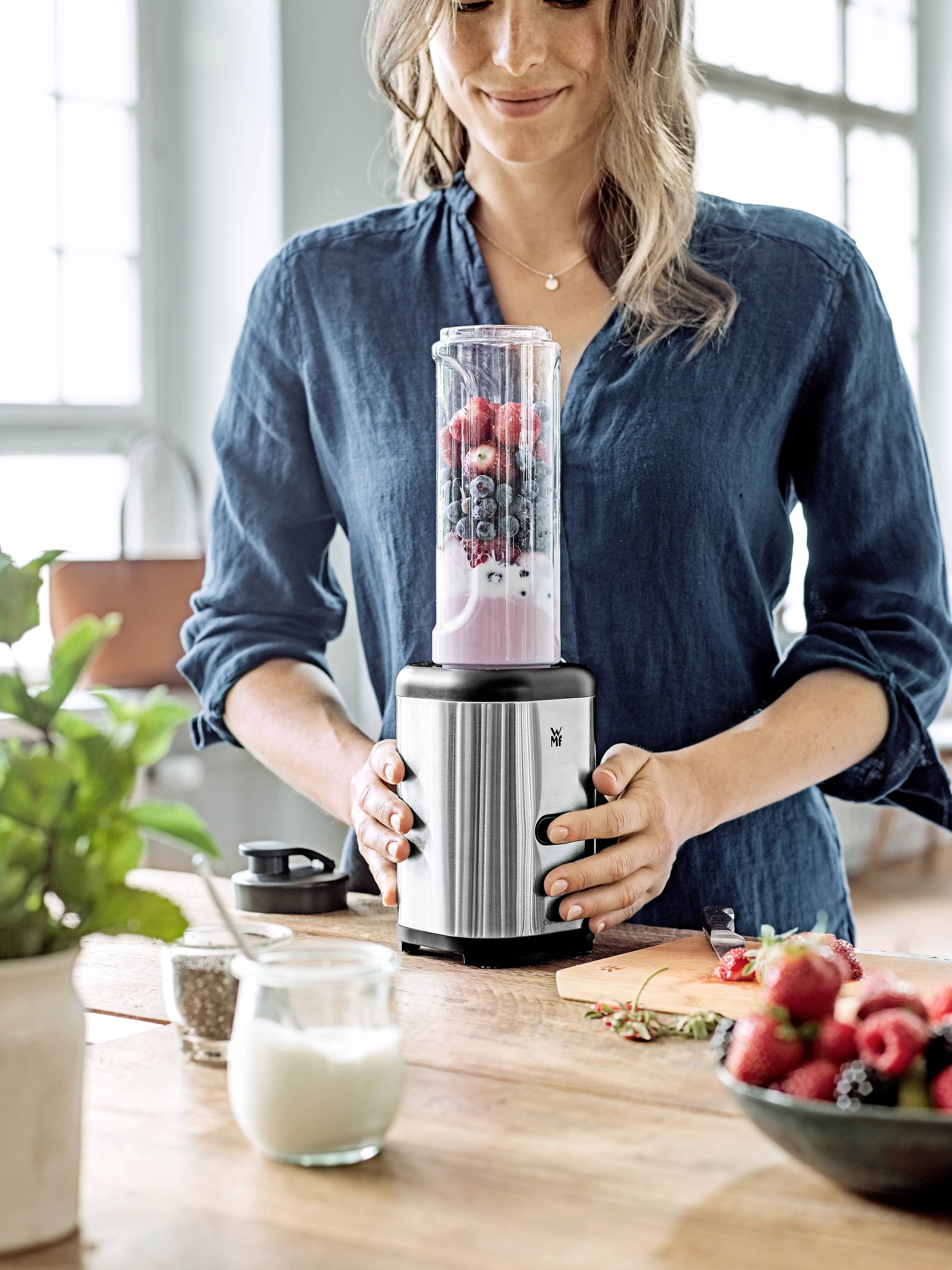 Donna con blusa blu che usa un frullatore per preparare uno smoothie di frutta. Bacche fresche e yogurt sul tavolo. Cucina sullo sfondo.