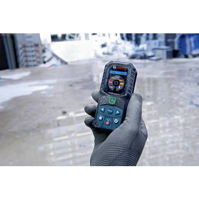 Una mano regge un telemetro laser in un cantiere edile. Lo strumento mostra i valori misurati, mentre sullo sfondo sono visibili materiali da costruzione sfocati.
