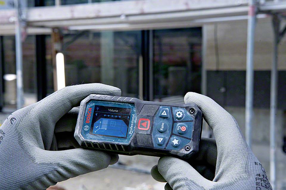 Una persona tiene un telemetro digitale con un ponteggio sullo sfondo. Il display mostra un diagramma per il calcolo del volume.