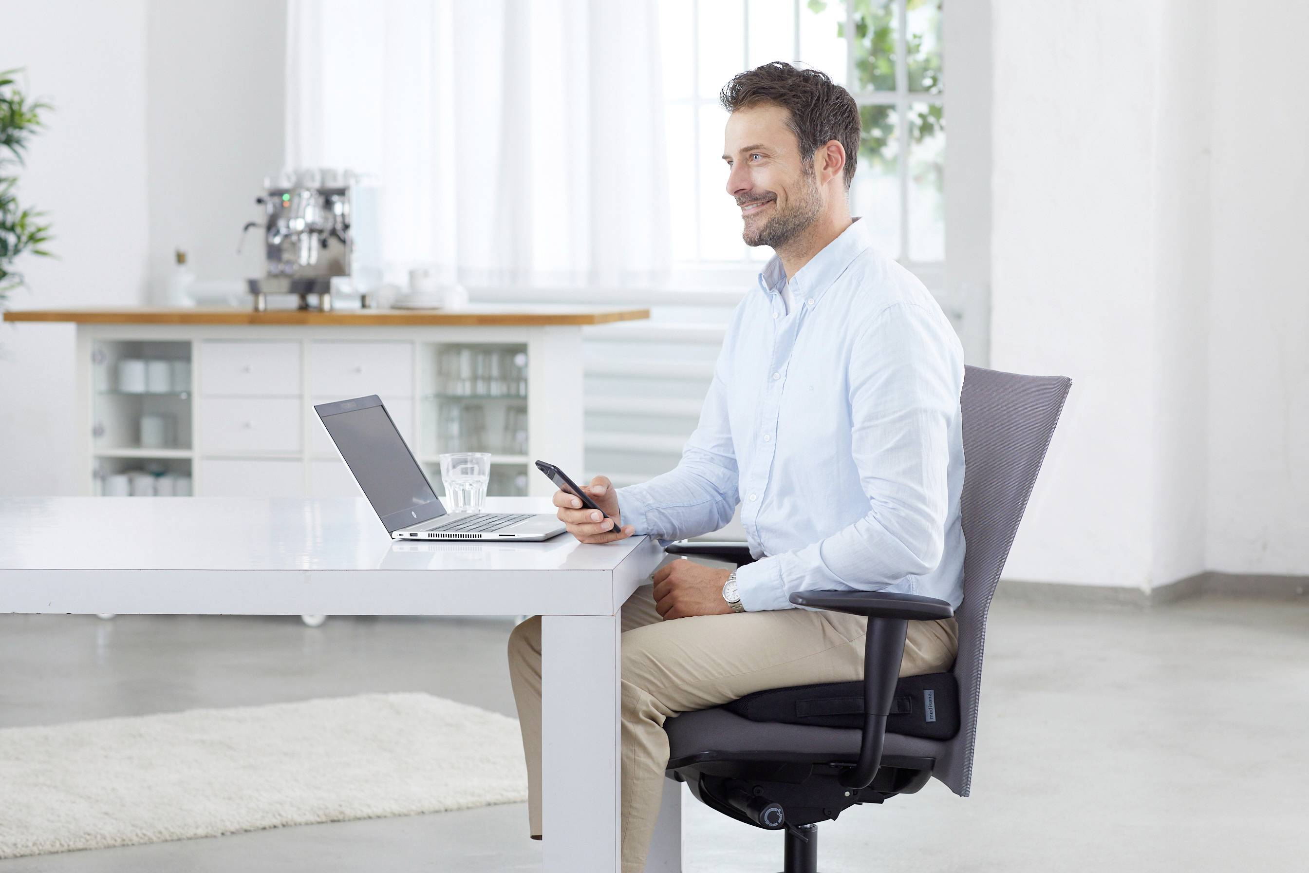 Un uomo seduto sorridente su una sedia da ufficio, davanti a un tavolo. Davanti a lui ci sono un laptop e un bicchiere d'acqua. Tiene in mano uno smartphone.
