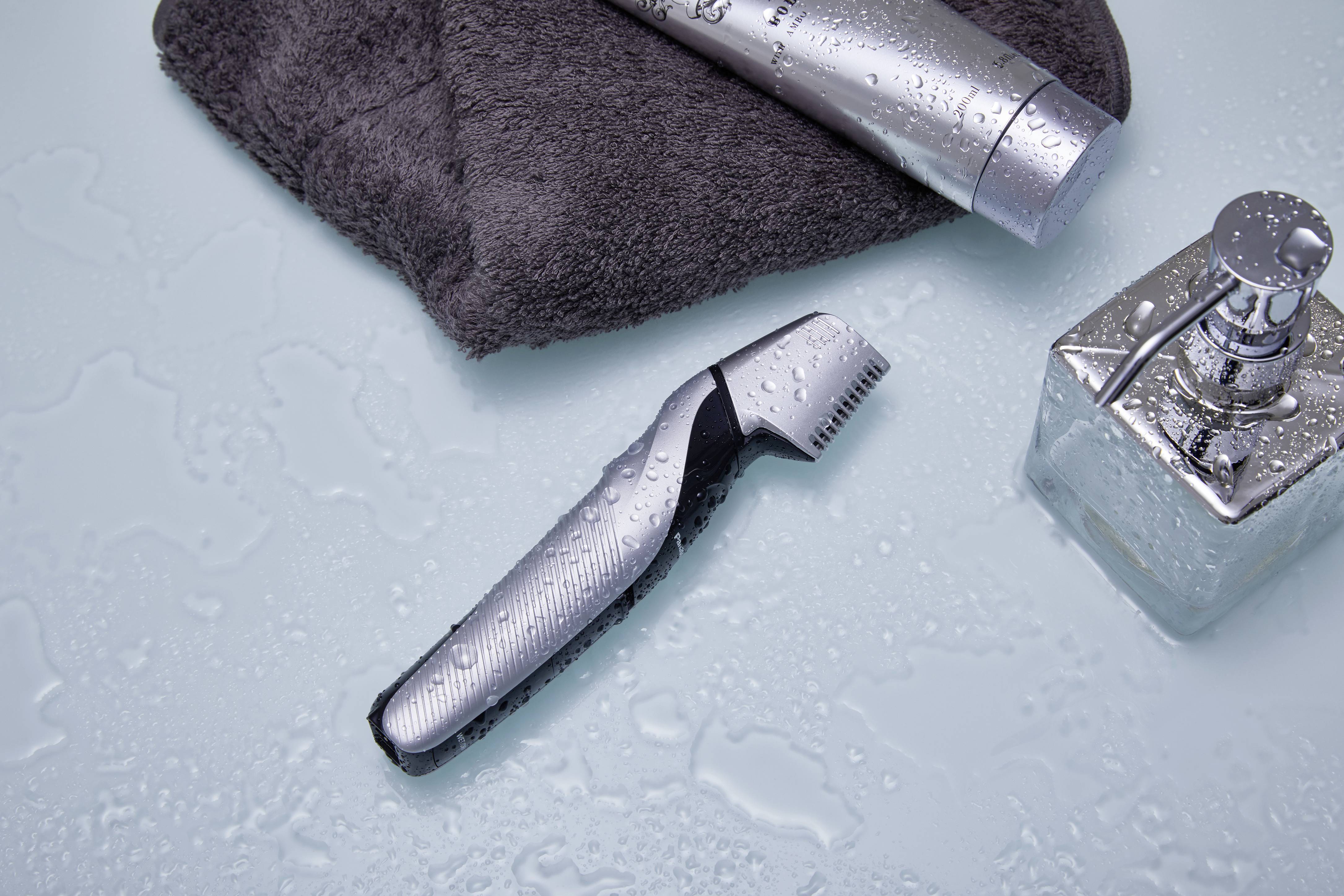 Un rasoio elettrico è posizionato su un tavolo bagnato, circondato da un asciugamano grigio, un tubetto di metallo e una bottiglia di vetro quadrata.