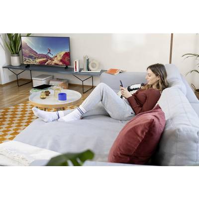 Una persona è seduta rilassata su un divano e guarda il proprio cellulare. Sullo sfondo, una televisione accesa mostra un paesaggio.