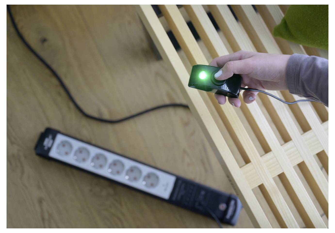 Una mano tiene un interruttore di alimentazione verde luminoso vicino a una struttura di letto in legno, controllando una ciabatta di alimentazione collegata a terra.
