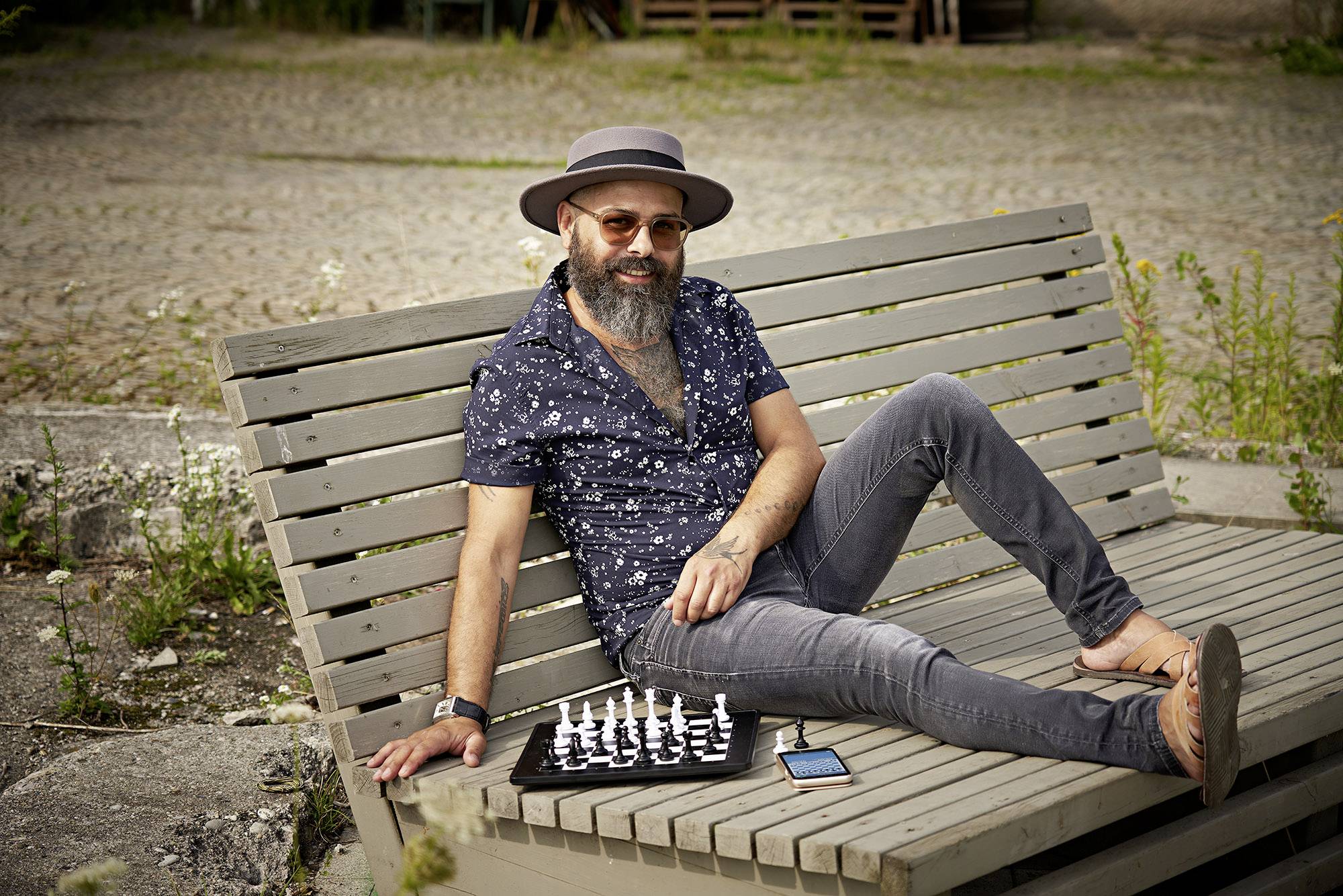 Un uomo con cappello e occhiali da sole è sdraiato sorridendo su una panchina all'aperto. Accanto a lui ci sono una scacchiera e un cellulare.